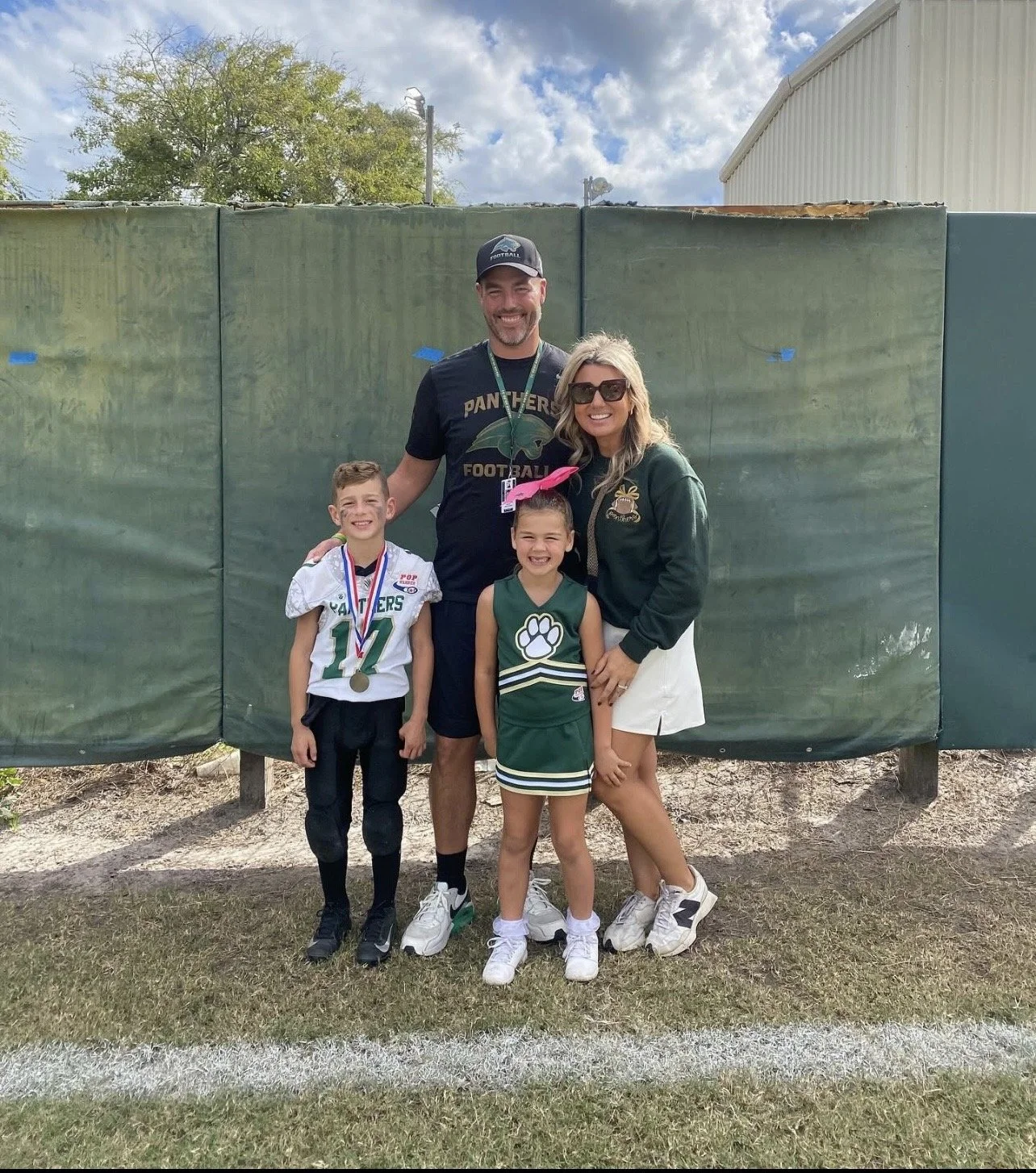 A family of four posing outdoors on a grass field, with a green privacy screen behind them. Two children are wearing sports jerseys, and the adults are standing with the children, all smiling.