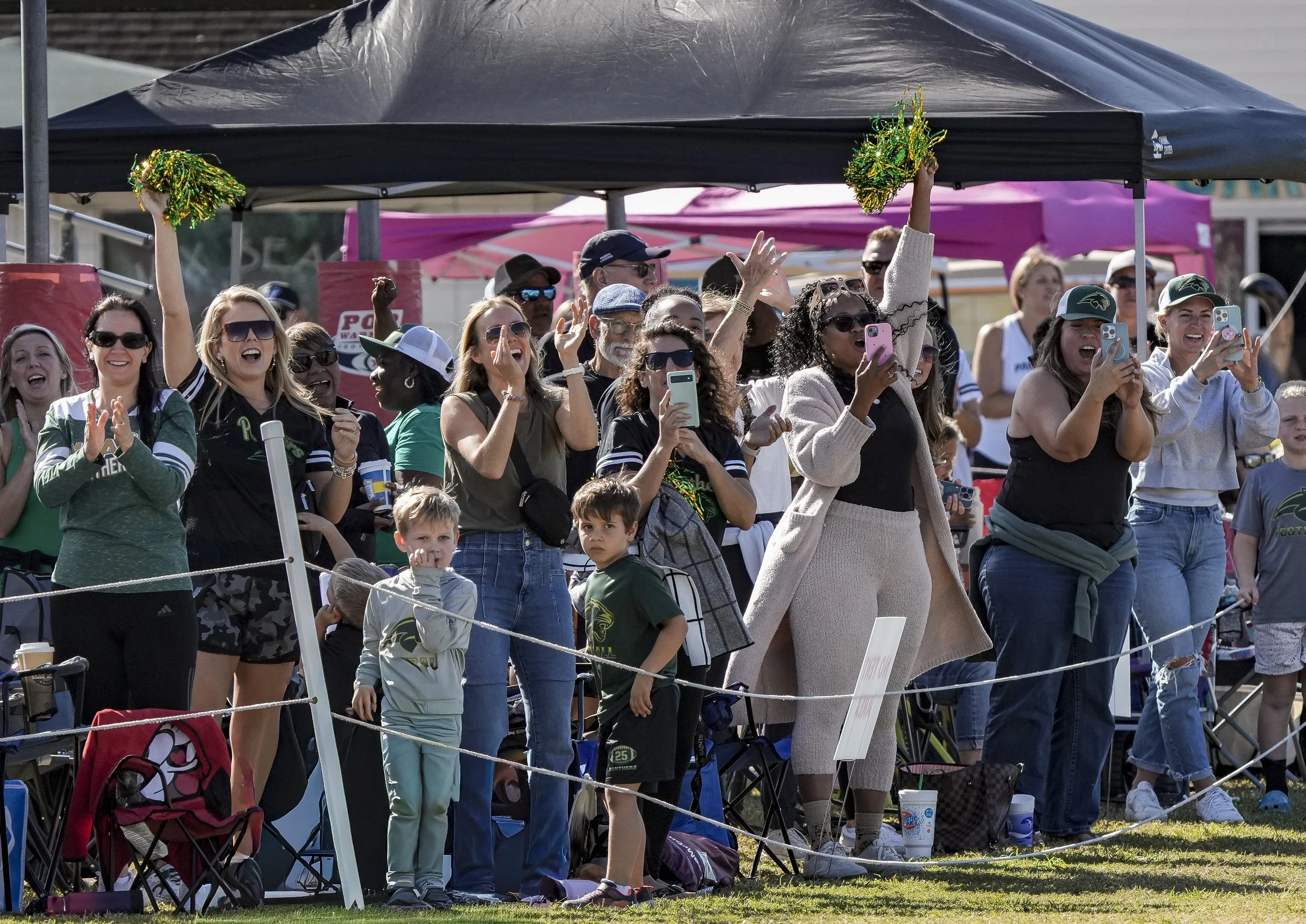 Crowd of people watching a sports event, some women cheering and taking photos, children standing in front, outdoor setting with tents in the background.