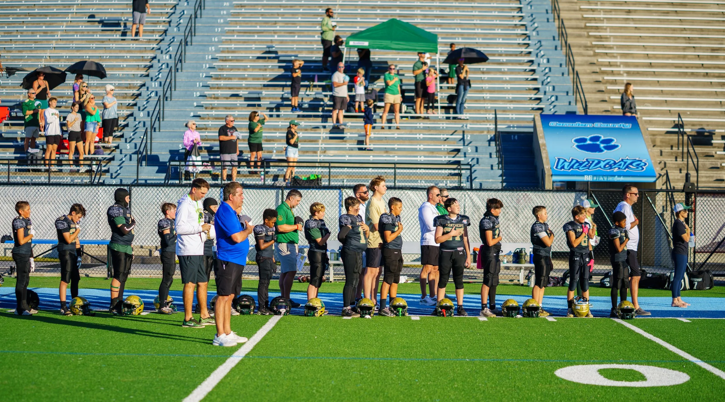 Young football players and coaches standing on the field with hands over their hearts during the national anthem at a stadium. The players are wearing black and gold uniforms, and helmets are placed on the ground in front of them. Spectators are seat
