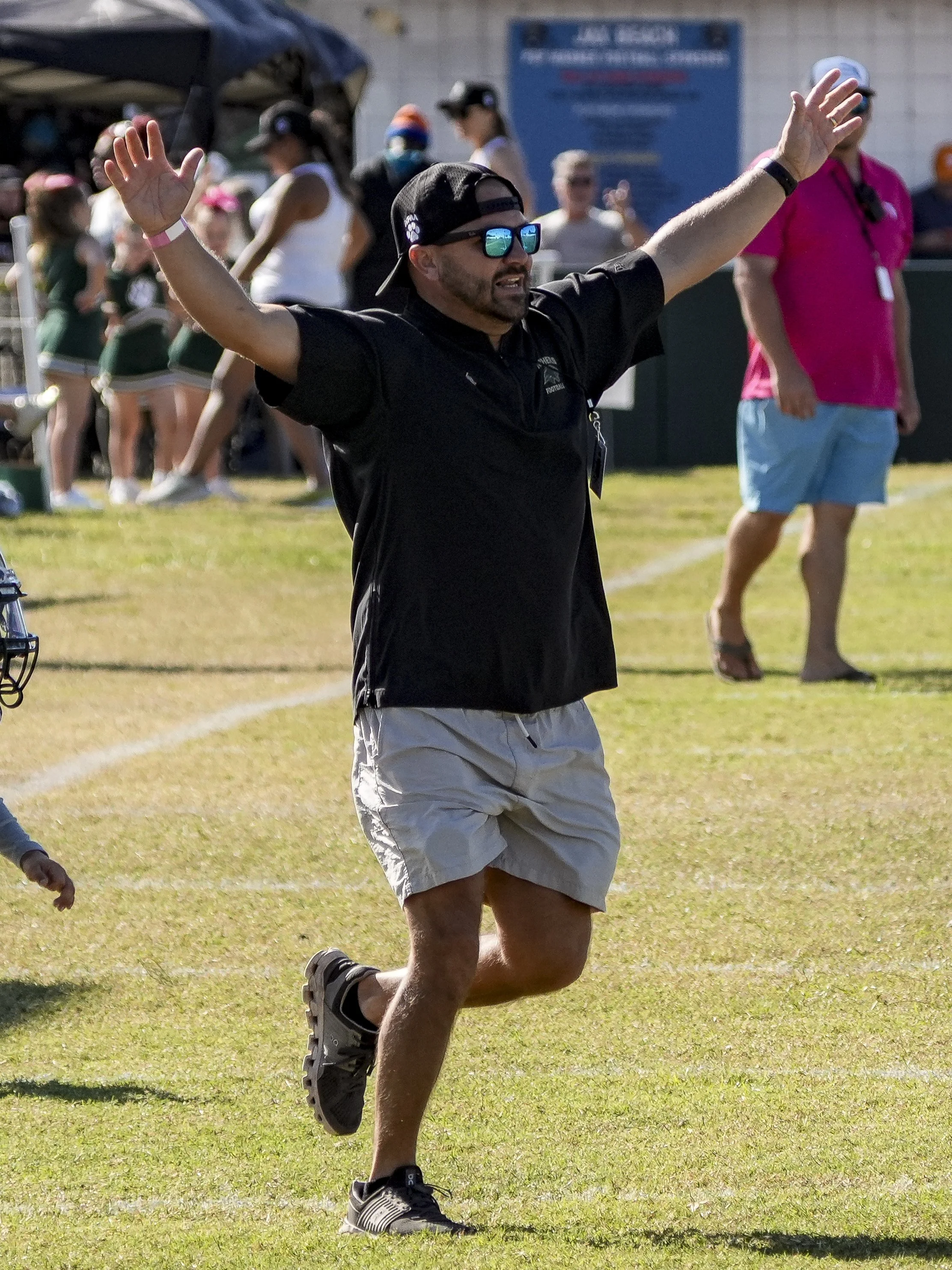 Man with sunglasses and black cap with arms raised, standing on a grassy field, with people in the background, possibly at a sporting event.