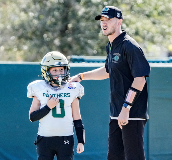 A young football player in a white Panthers jersey and helmet listening to a coach during a game or practice outdoors.