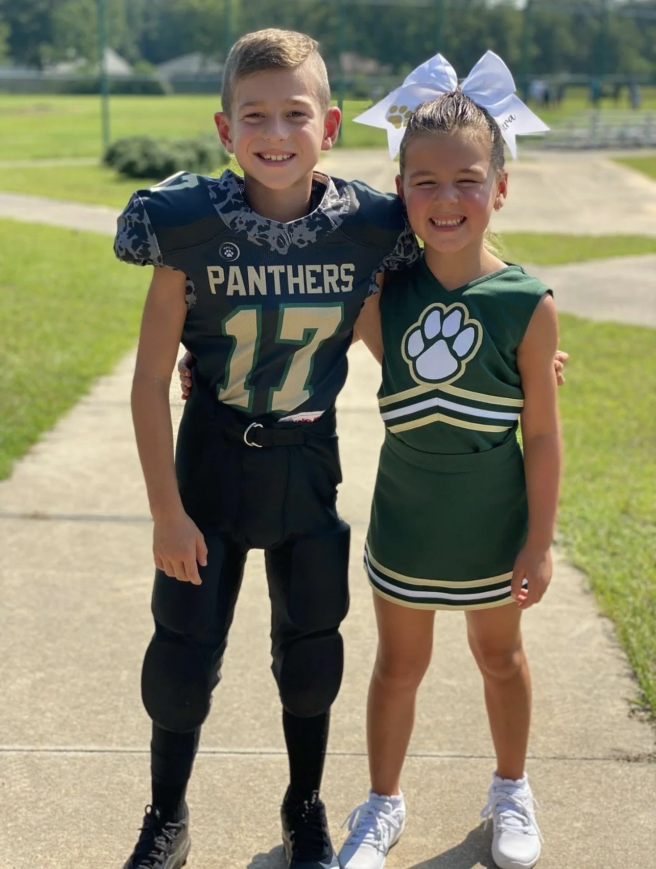 A boy in a football uniform and a girl in a cheerleading outfit standing together outdoors on a sidewalk, smiling.