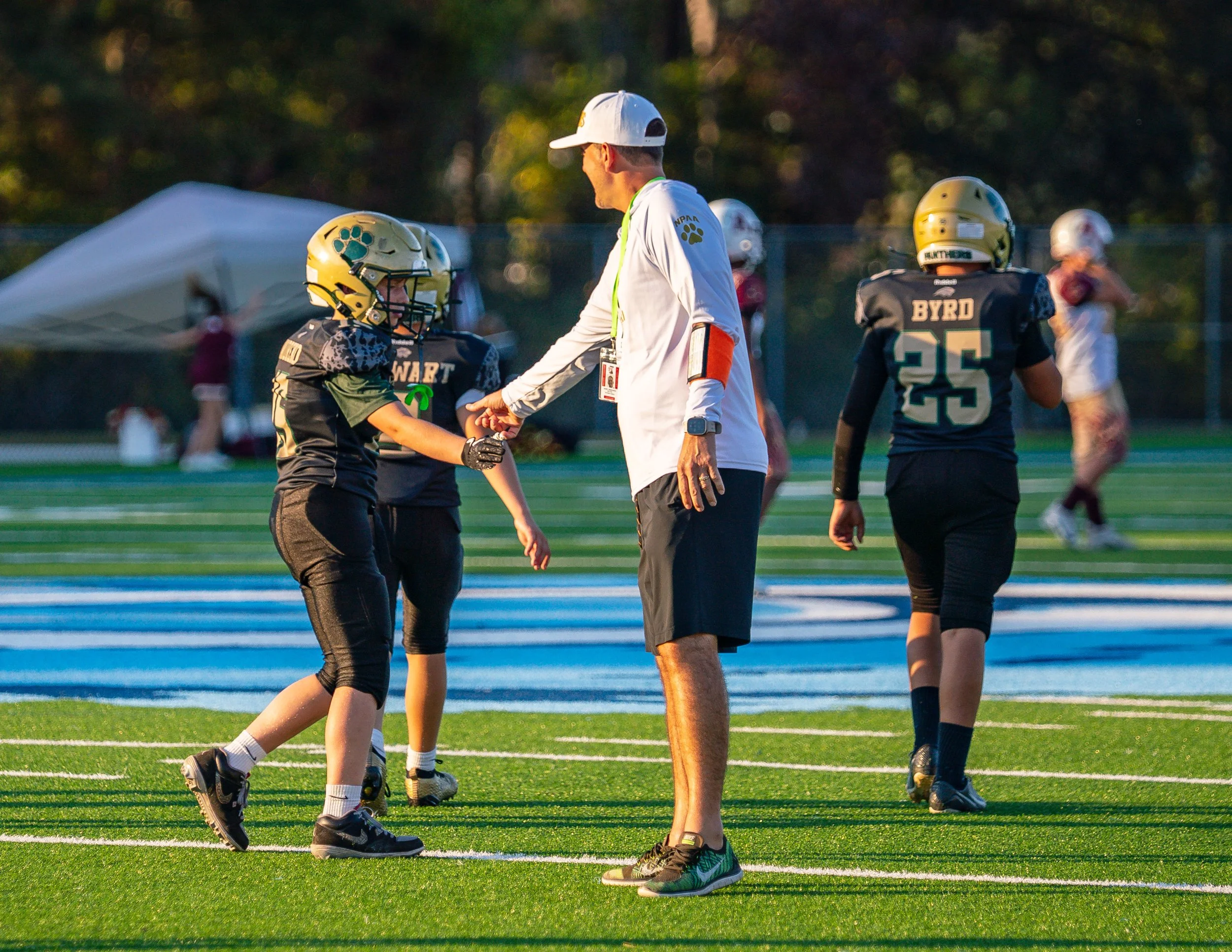 A youth football coach shaking hands with a young player on the field, with other players and spectators in the background at a football game.