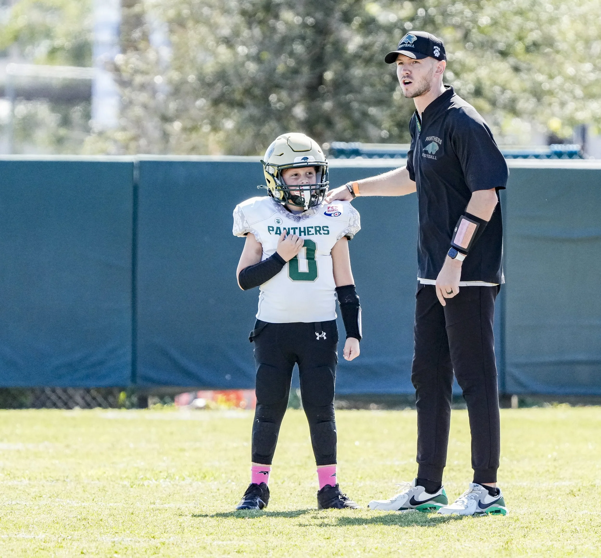 A young football player in a white jersey with green and black accents, wearing a helmet and pads, standing on a football field next to a coach, who is dressed in a black shirt and pants with a cap. The coach appears to be giving instructions to the 