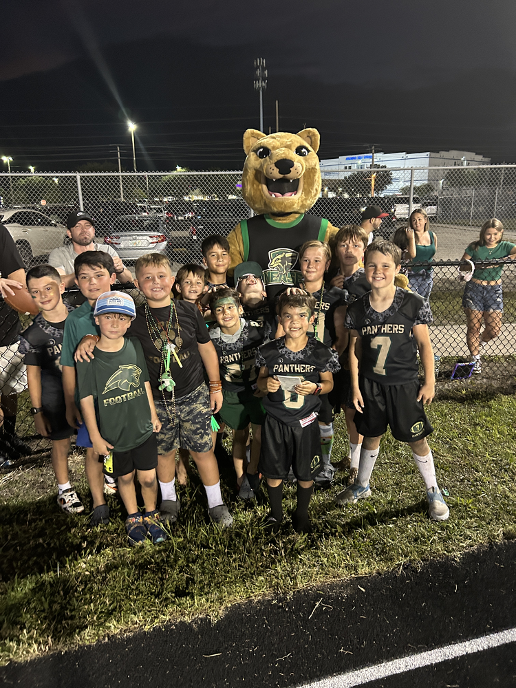 A group of young children in football uniforms posing with a mascot in a lion costume at night near a wire fence, with a parking lot and stadium lights in the background.