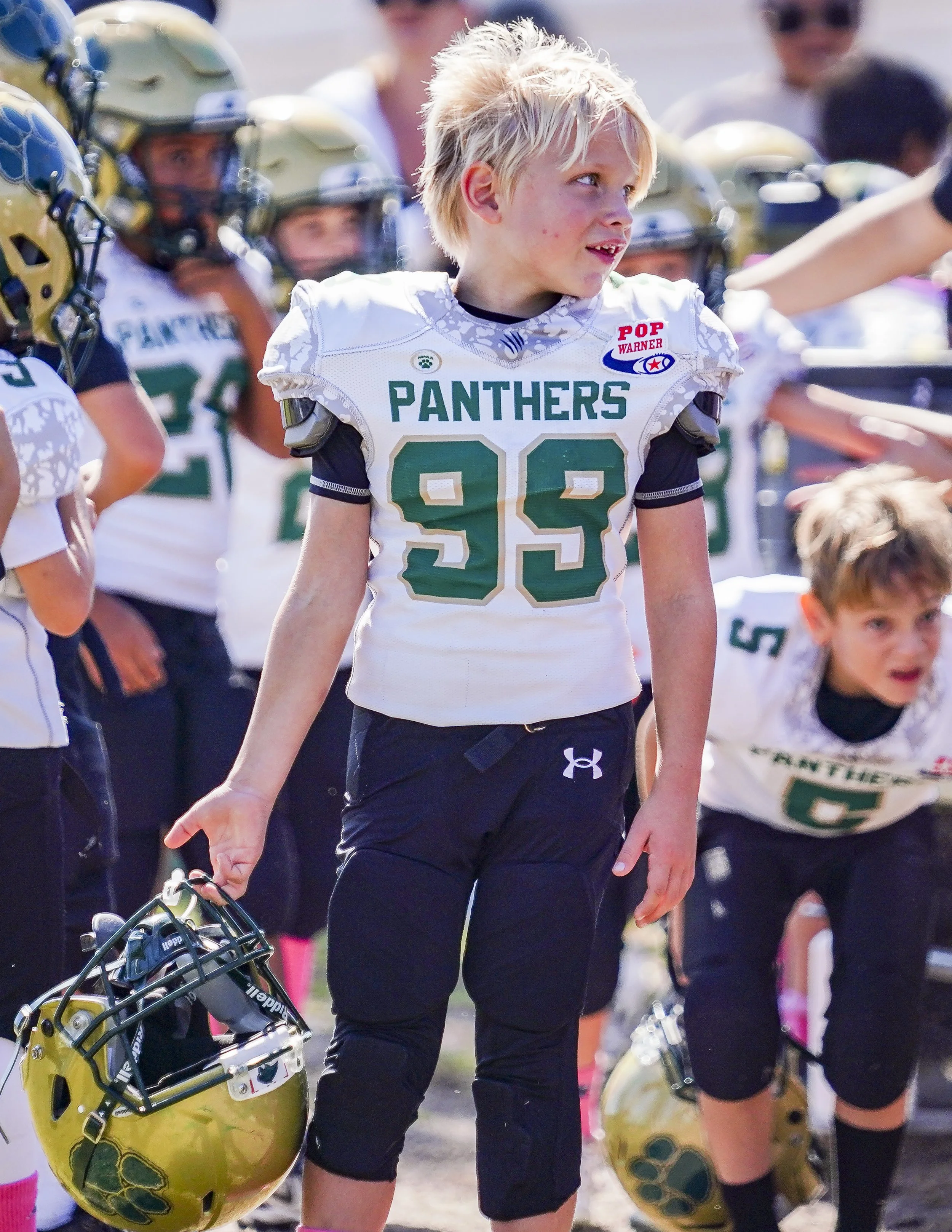A young boy in a football uniform holding a helmet, standing among other kids in similar uniforms on a football field.