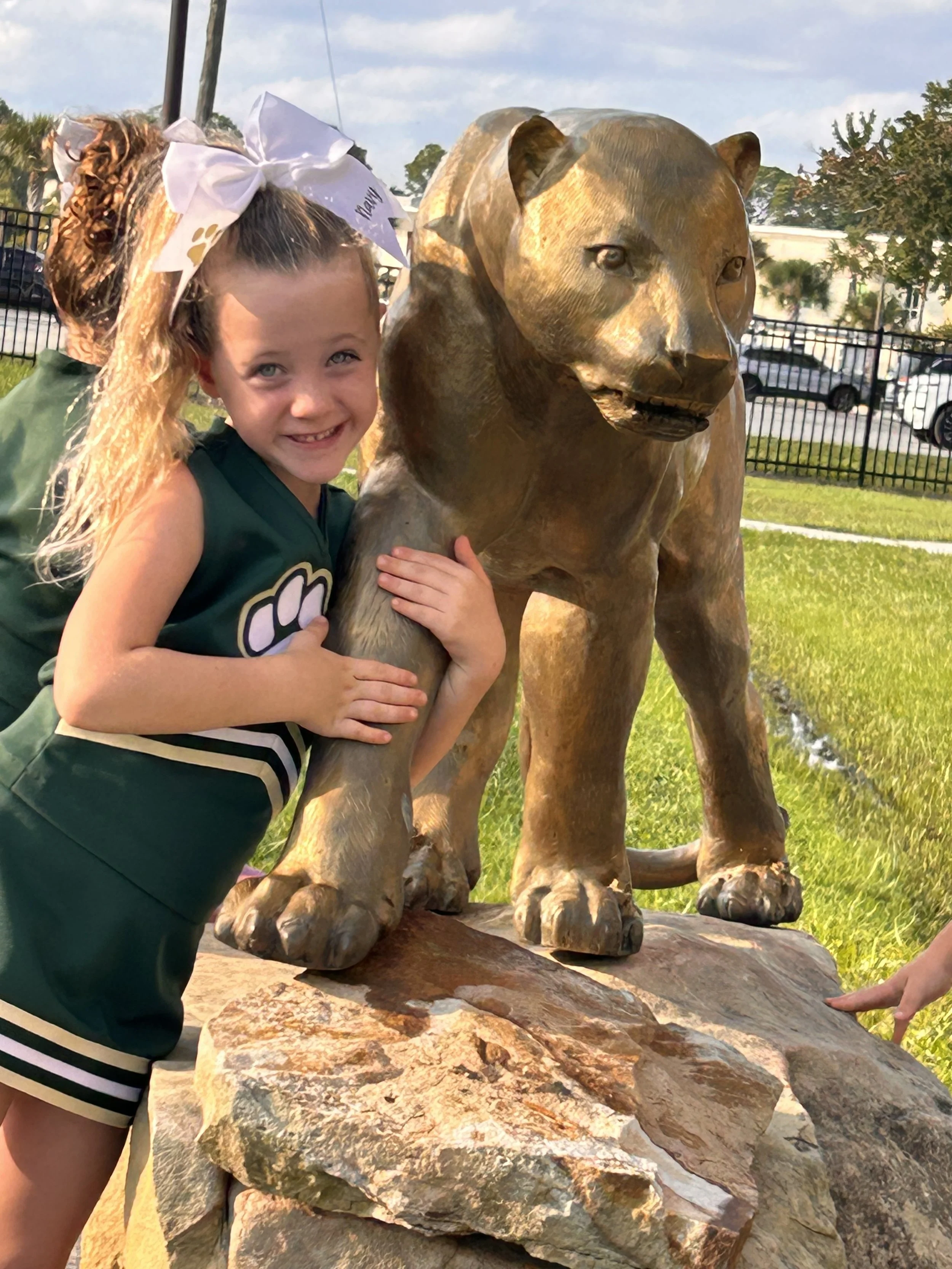 A young girl wearing a green cheerleading outfit with a white bow in her hair smiles while hugging a bronze lion statue outdoors on a sunny day.