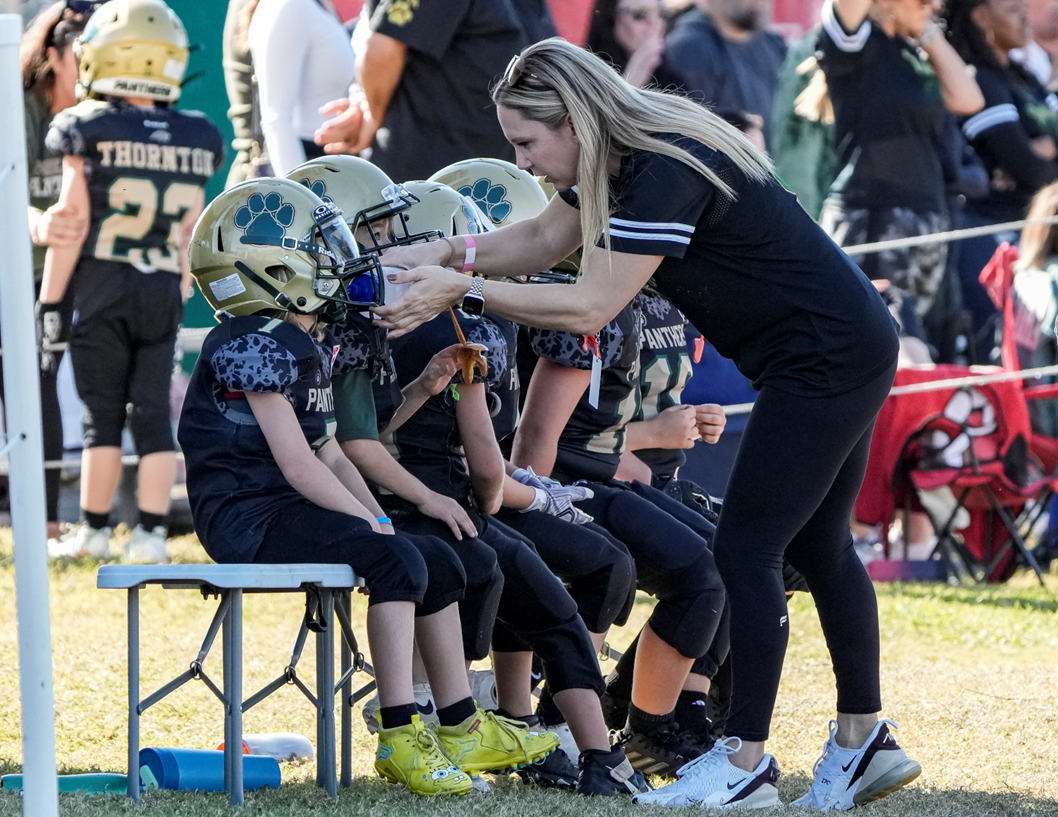 A woman helping young football players with their helmets on the sidelines of a game.