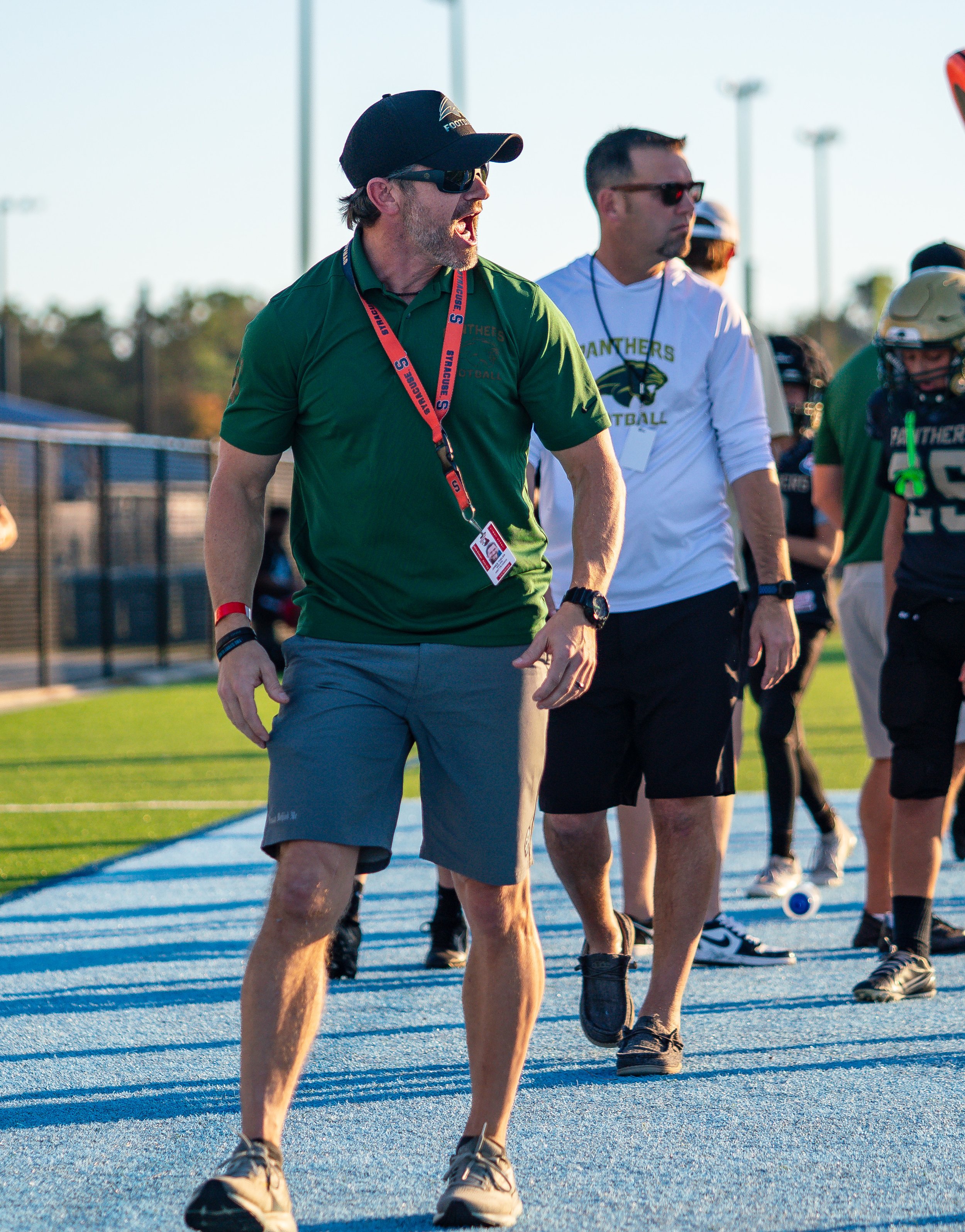 Football coach with sunglasses and whistle on sidelines during game, wearing green shirt and shorts, standing on blue track with team players nearby.