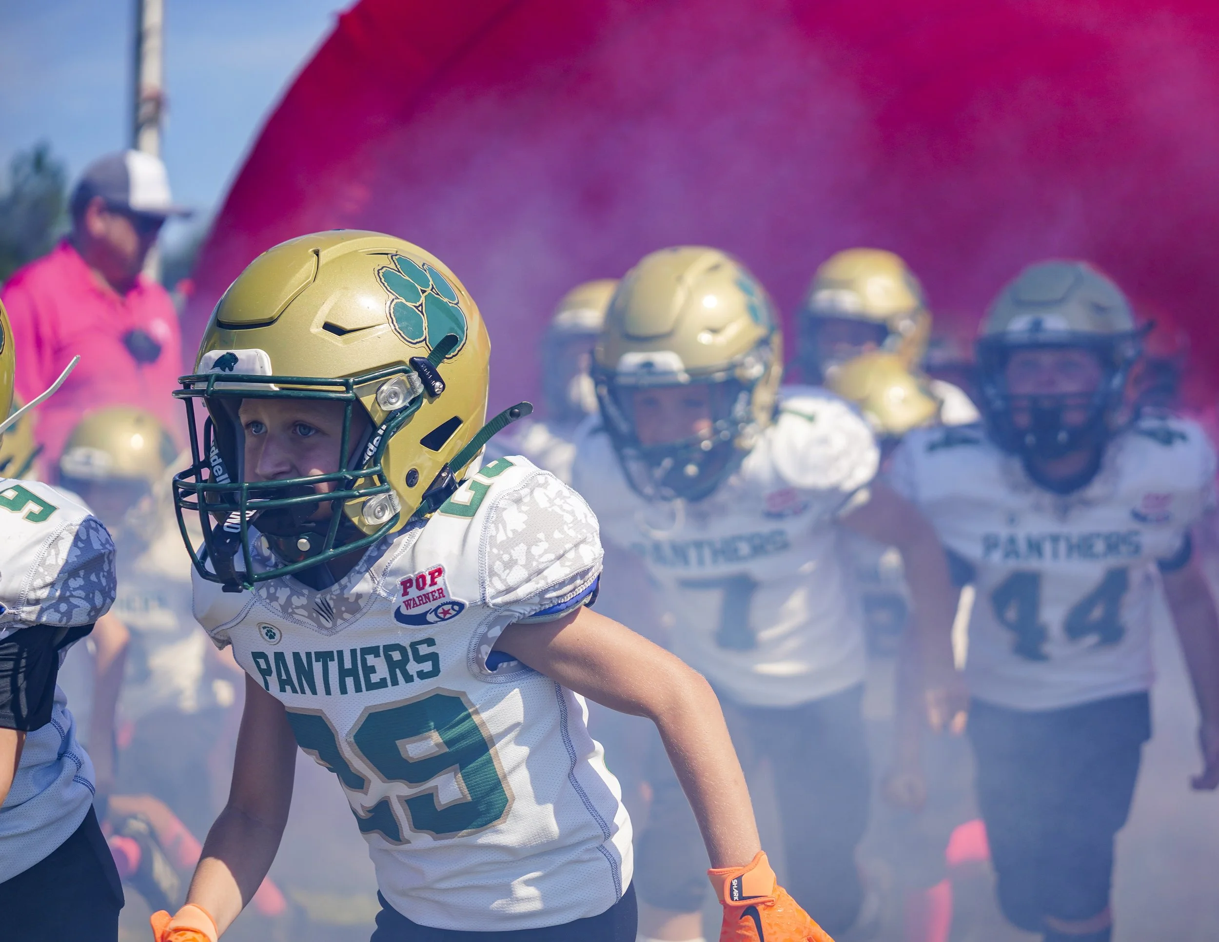 Youth football team called Panthers, with kids in white jerseys and gold helmets, running onto field through a cloud of pink smoke during a game or practice.