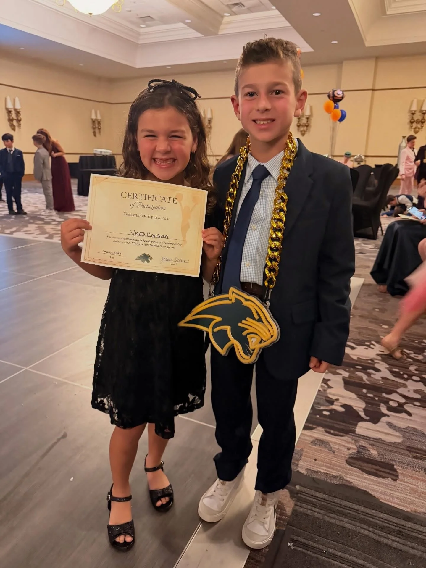 A young girl holding a certificate of participation and a boy wearing a suit with a football helmet necklace at an indoor event.