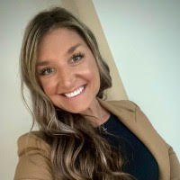 A woman smiling and taking a selfie indoors, with light-colored walls in the background.