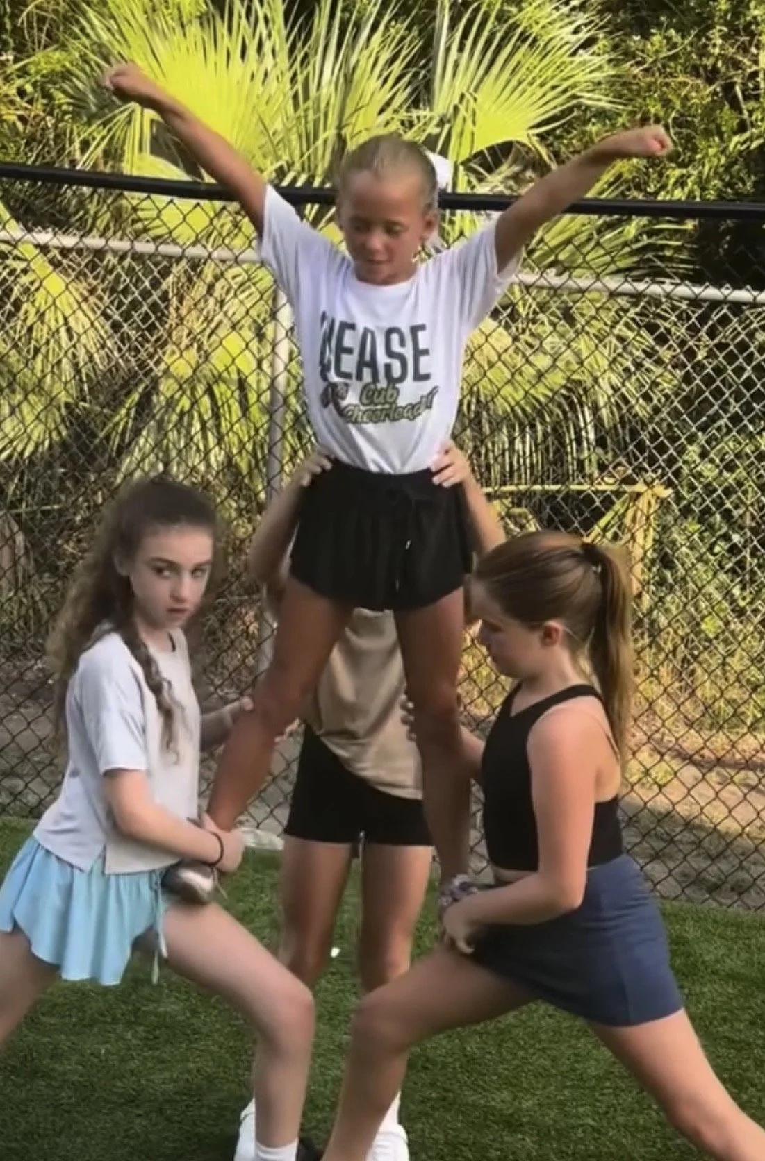 Three young girls in sports clothes forming a human pyramid outdoors, with a girl standing on the shoulders of two others. The girl standing has her arms raised and is smiling, while the other two hold her ankles. There is a chain-link fence and gree