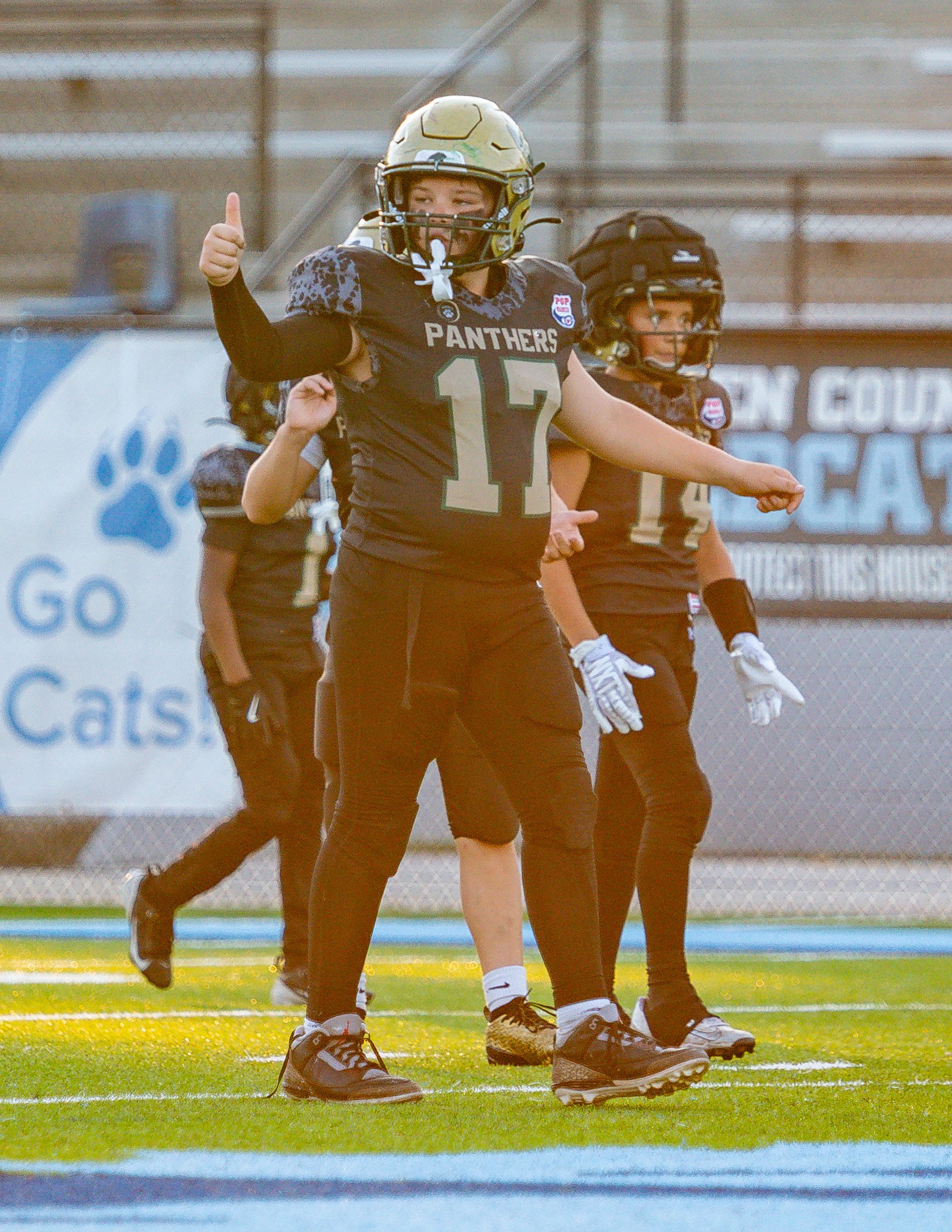 Young female football player in a black uniform with the number 17, wearing a gold helmet, giving a thumbs up on the field during a game.