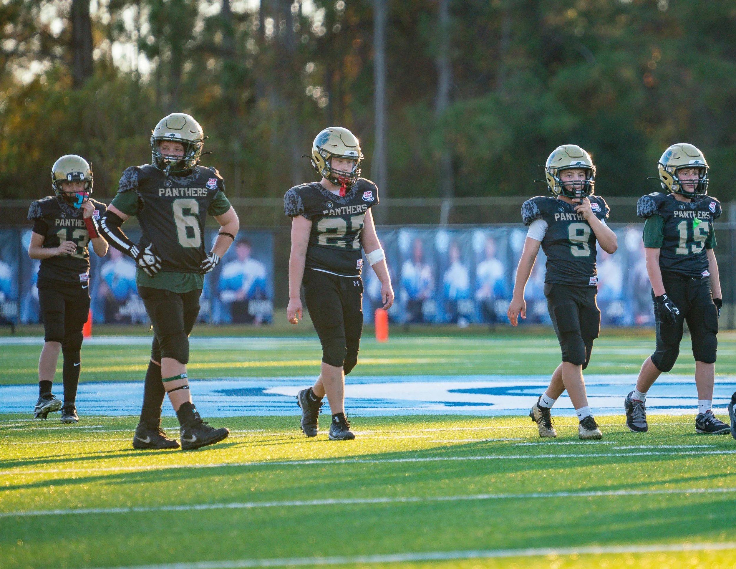Youth football players in black and green uniforms walking on the field during sunset, with a forest background and bleachers with photos of players.