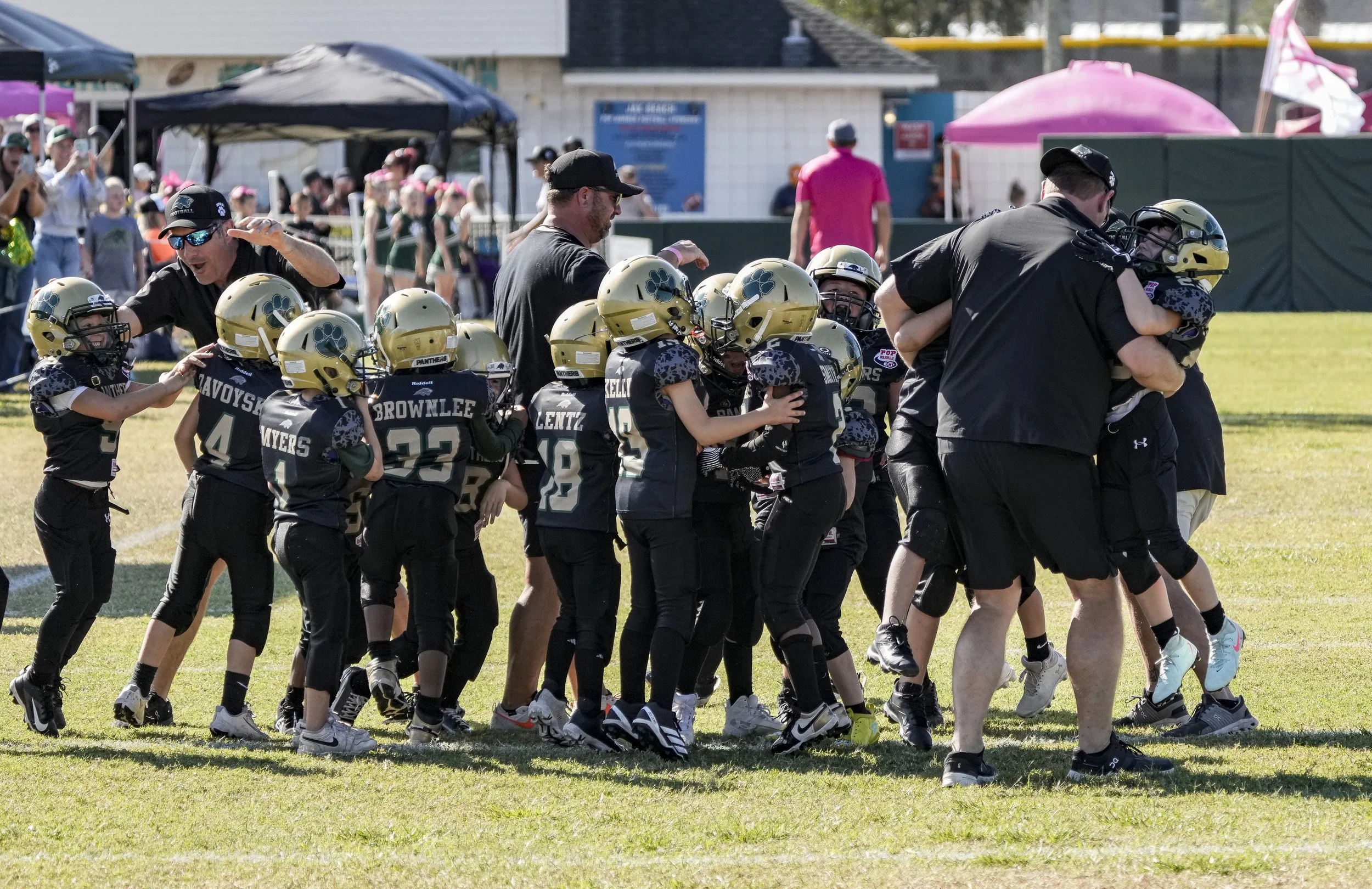 Youth football players in black and gold uniforms celebrating with coaches on a grassy field during a game or practice.
