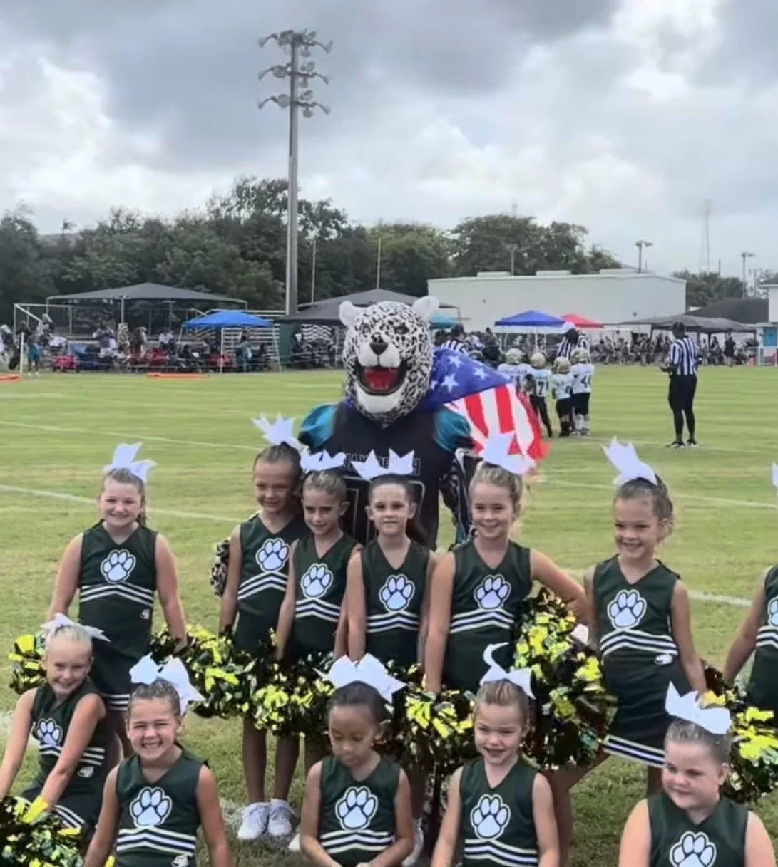 Group of young cheerleaders in green uniforms with paw print logos, posing in front of a jaguar mascot draped in an American flag at a youth football game on a grassy field under a cloudy sky.