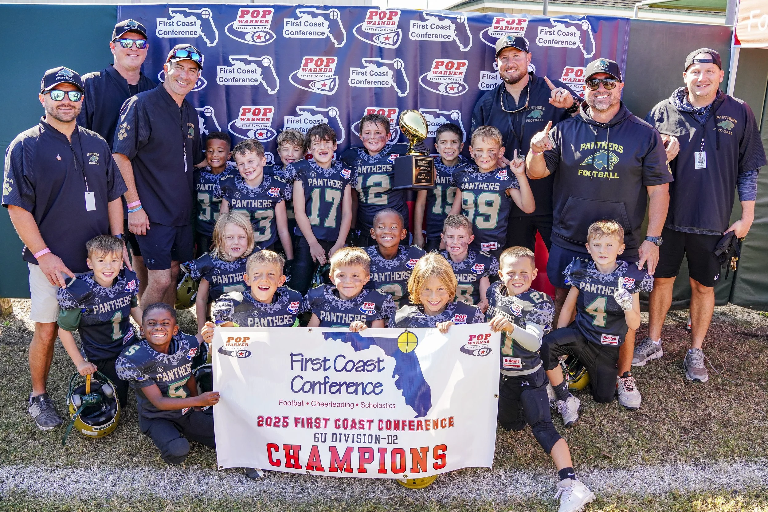 Youth football team called the Panthers with coaches and staff, celebrating as 2025 First Coast Conference champions, holding a trophy, in front of a backdrop with logos.