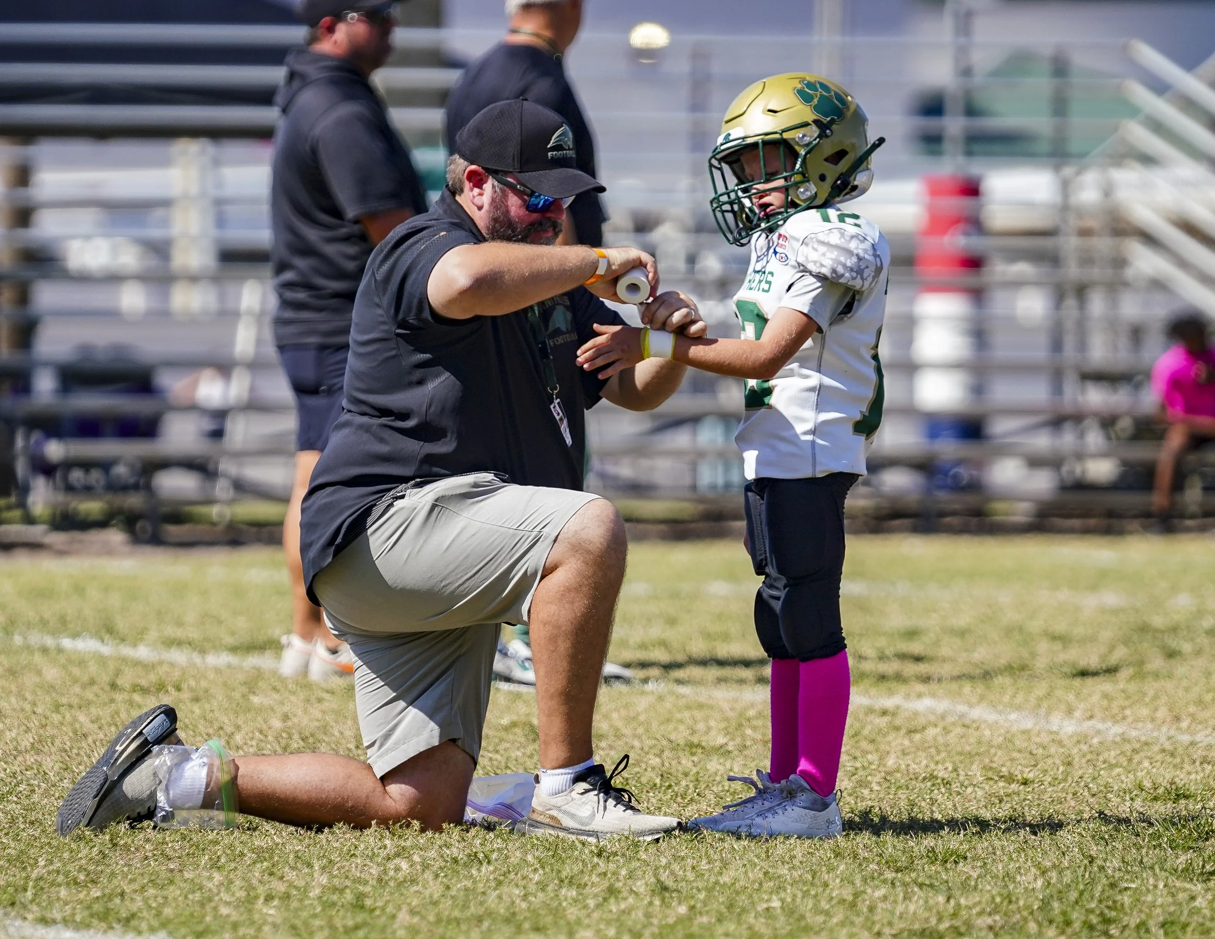 A young football player wearing a white jersey with green and gold accents, black pants, pink socks, and a gold helmet, is receiving medical attention from a coach on the field during a game.
