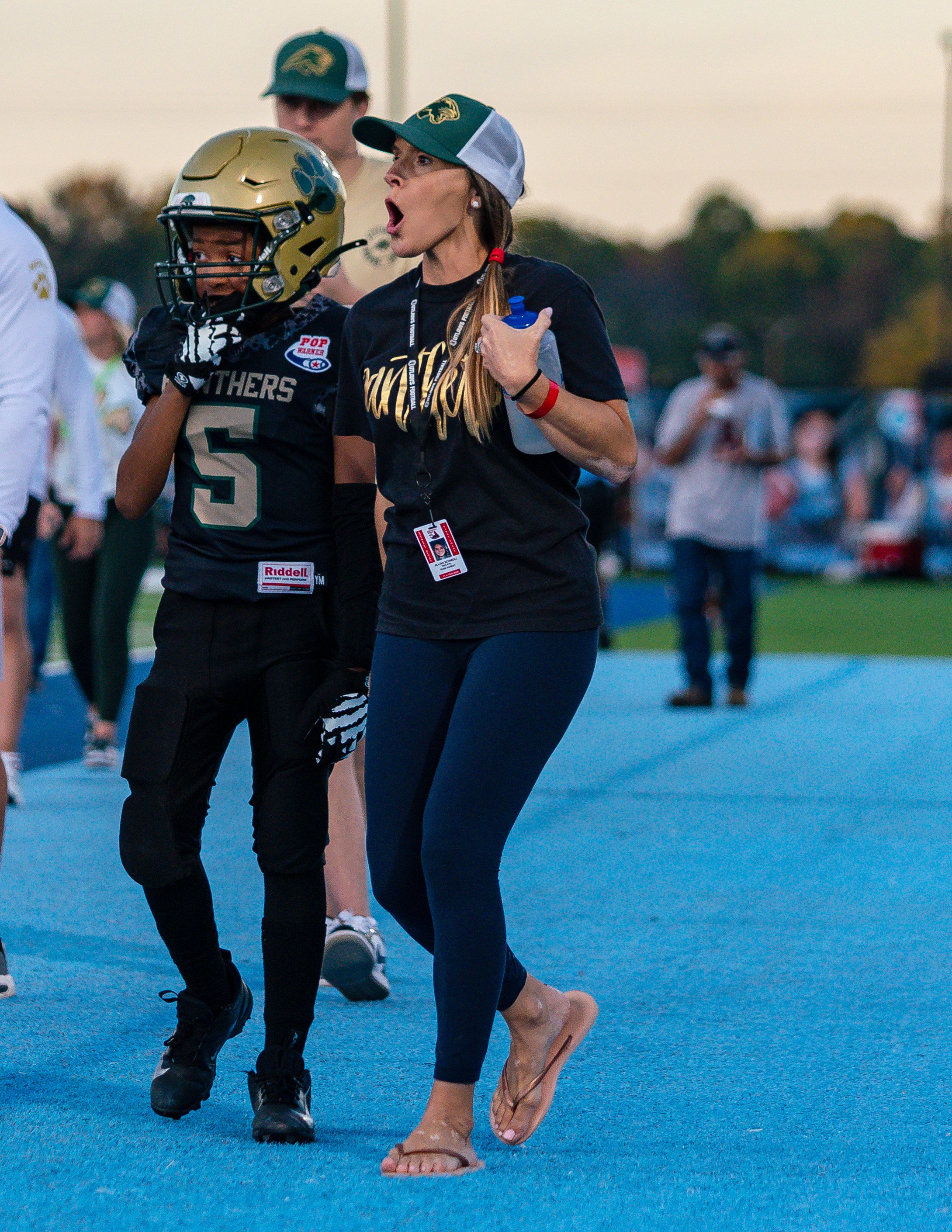 A woman with a ponytail wearing a cap, black T-shirt, leggings, and flip-flops, holding a water bottle, walking on a blue field during a sports event. A young football player wearing a helmet and uniform with the number 5 stands nearby, with several 