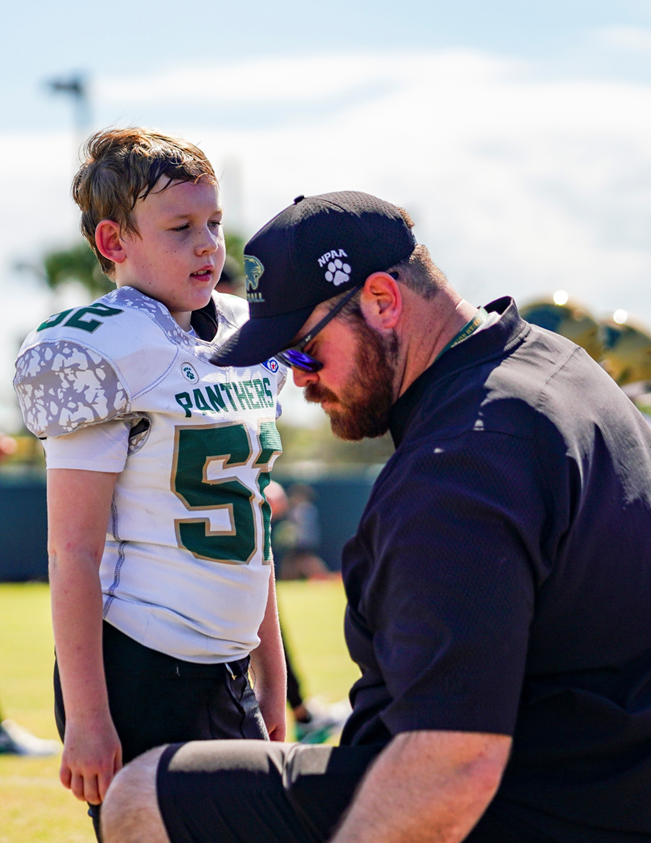 A young boy in a football uniform is talking to a coach on the field during a game or practice. The boy appears attentive, and the coach is kneeling in front of him, wearing sunglasses and a cap.