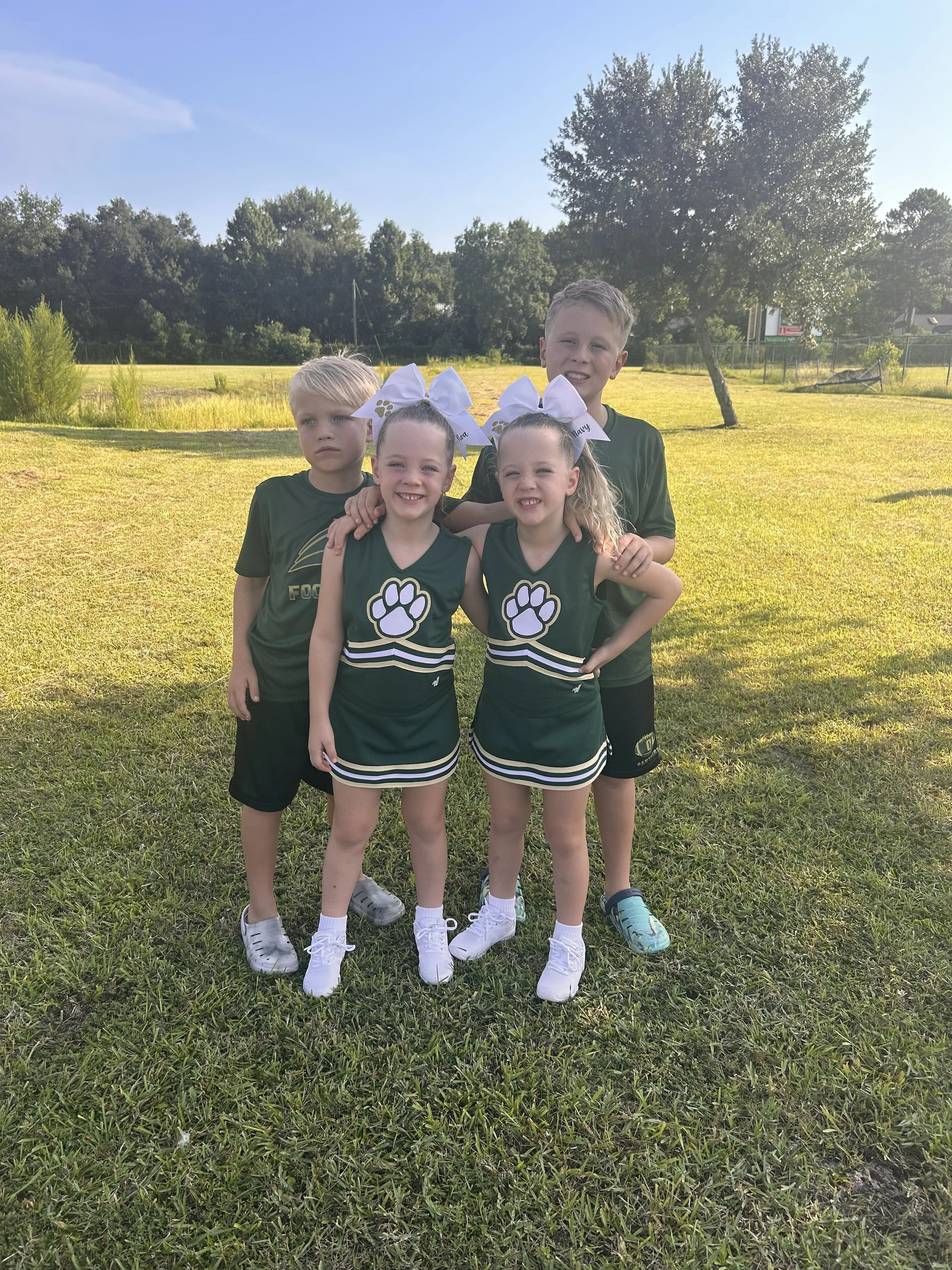 Four children in cheerleading uniforms with green and black colors and paw print logos, standing on grass with trees and a sunny sky in the background.