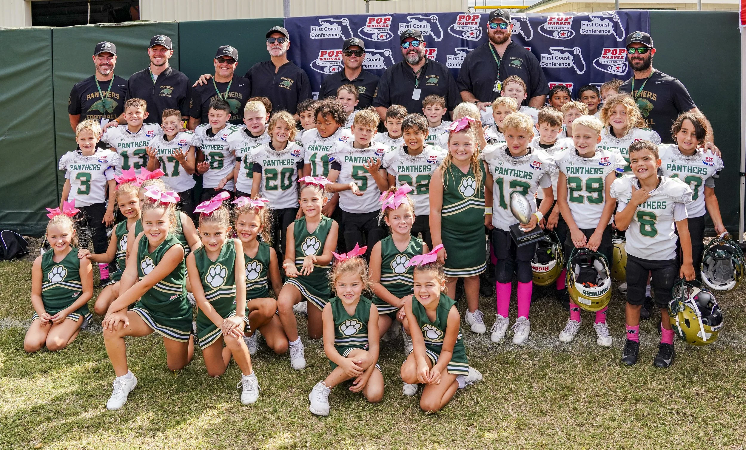 A large group of young children, coaches, and cheerleaders posing together on a football field after a game or event, with a backdrop featuring sponsors and team logos.