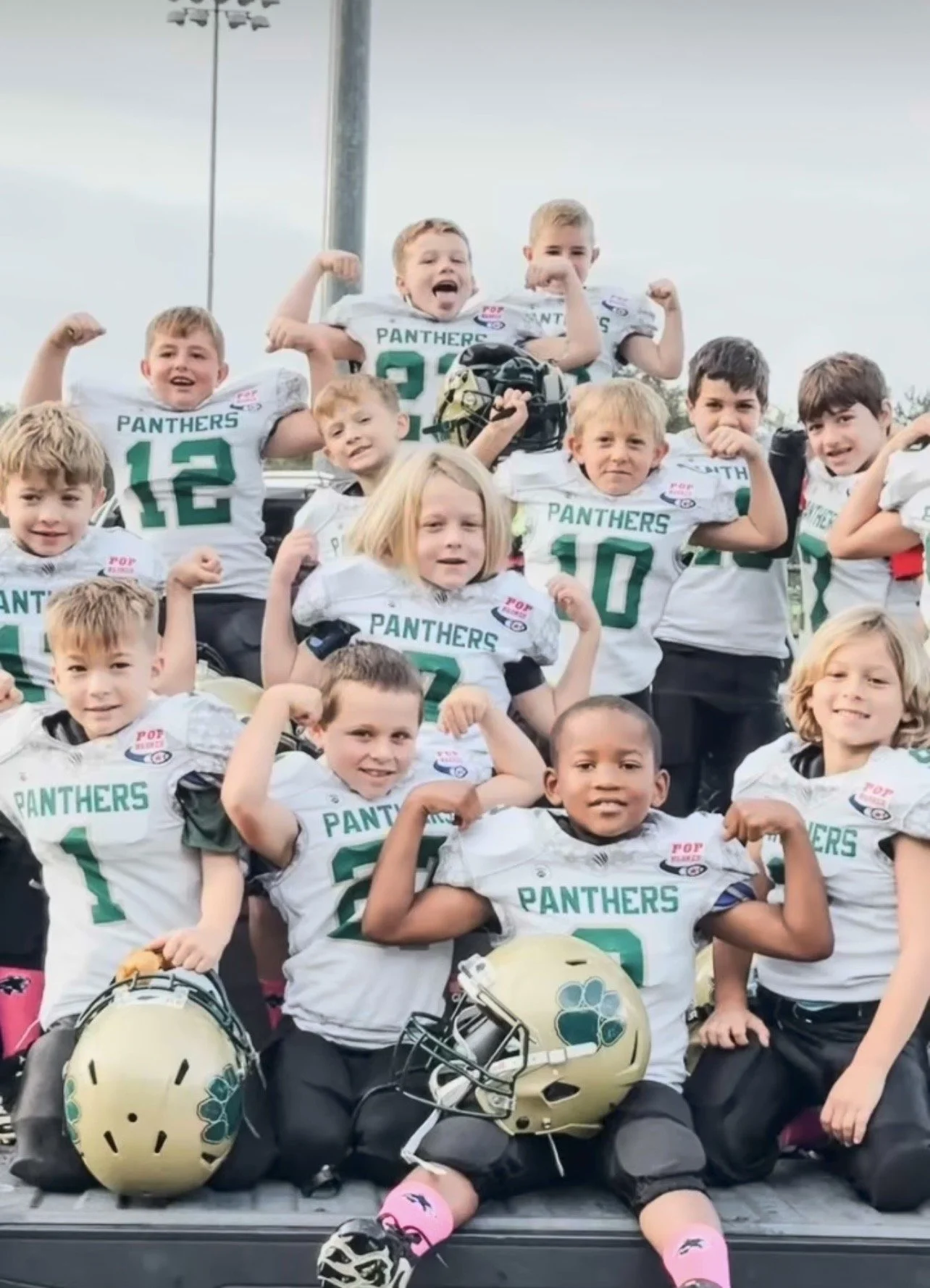 A group of young children in football uniforms, some flexing their muscles and showing their biceps, posing for a team photo on a football field. They are wearing white jerseys with 'PANTHERS' printed on the front and football helmets placed in front