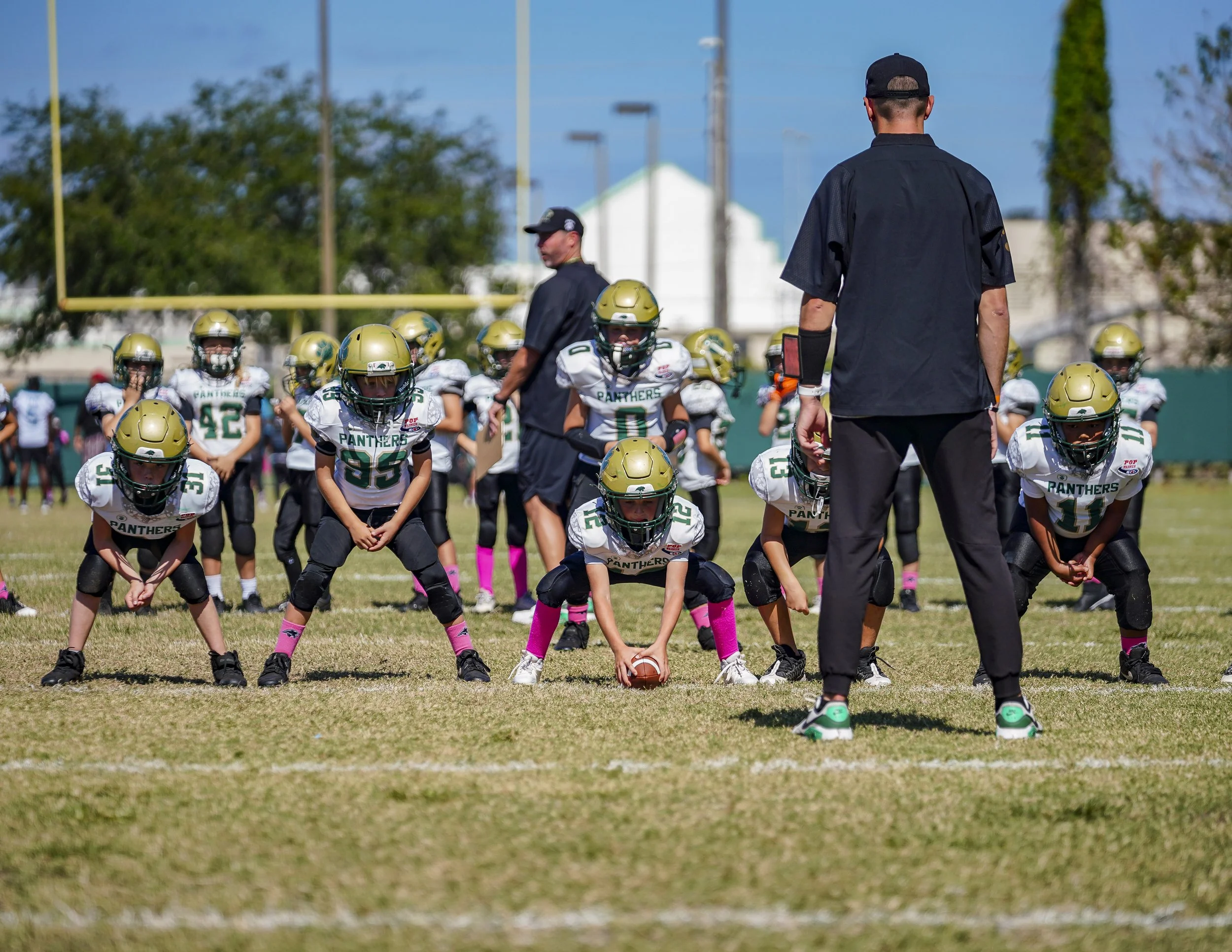 Youth football team in white jerseys, black pants, and gold helmets practicing on a field with coaches standing in front
