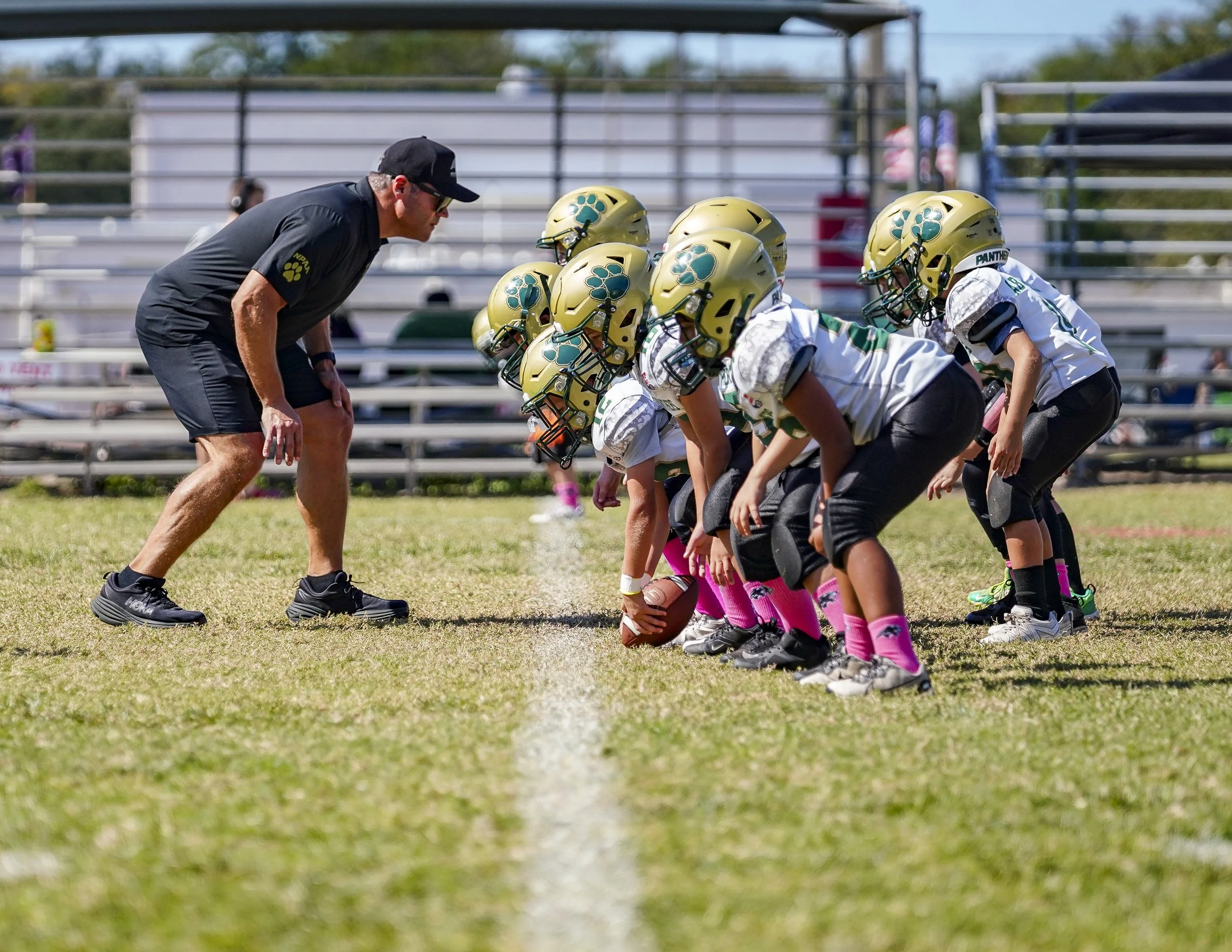 Youth football team in white jerseys and gold helmets practicing on field with coach.