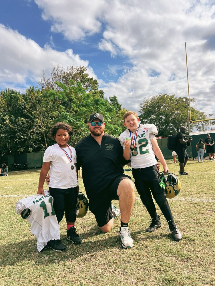 A football coach kneeling on the field with two young players, one holding a helmet, after a game.