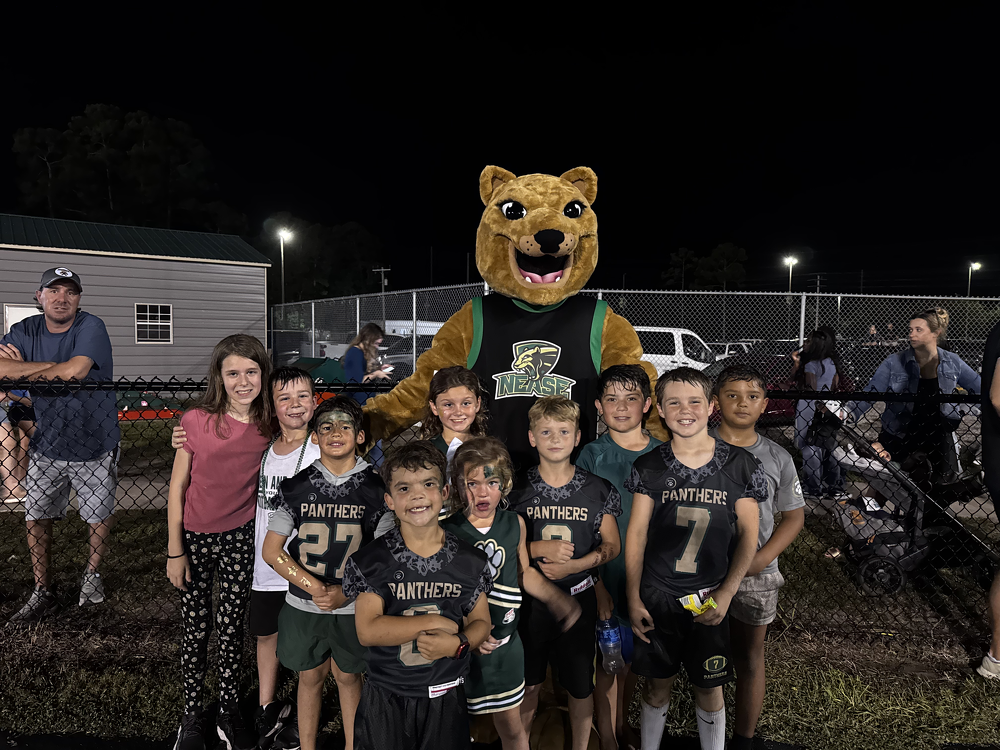 A group of children in football uniforms standing in front of a fence with a mascot dressed as a large panther at a nighttime outdoor sports event.