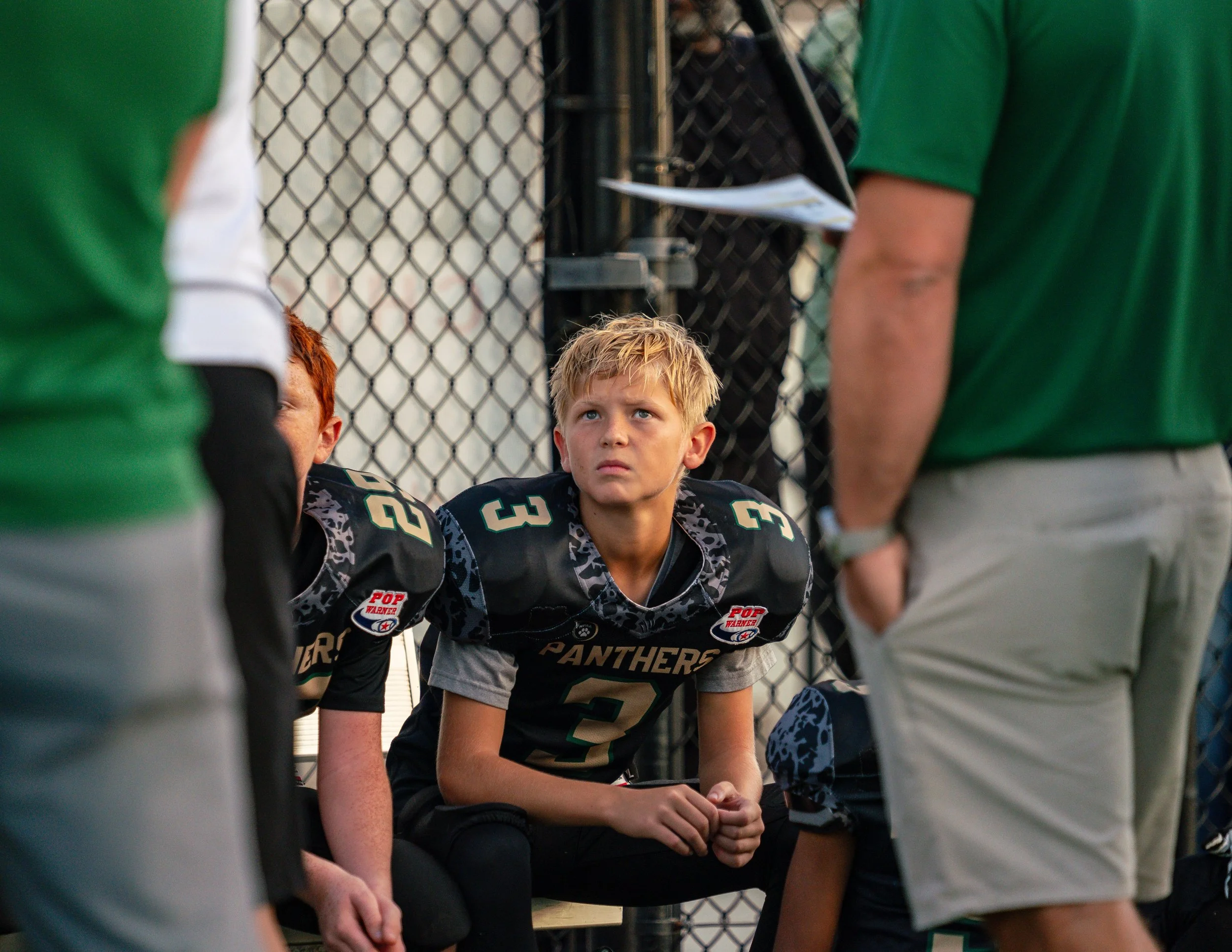 Young football player in black and gray uniform sitting on the ground, attentively listening during a timeout or team meeting, with coaches standing nearby, at a sports field enclosed by a chain-link fence.