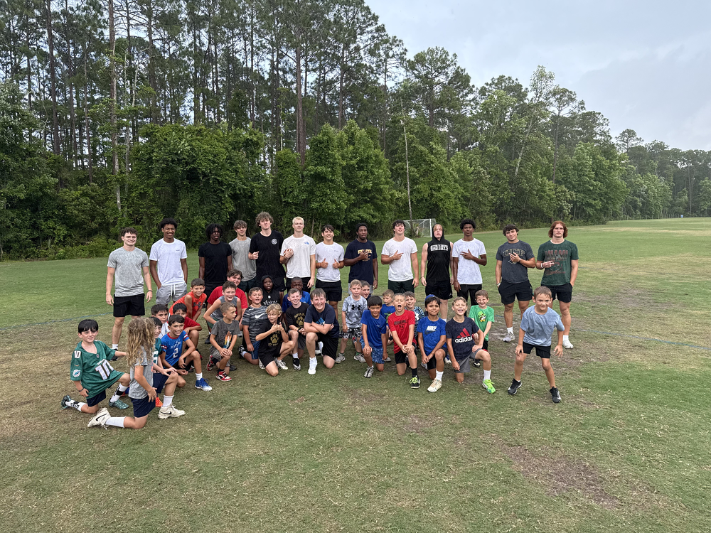 Group of children and teenagers on a grassy field, some kneeling and some standing, with a wooded area in the background, during daytime.