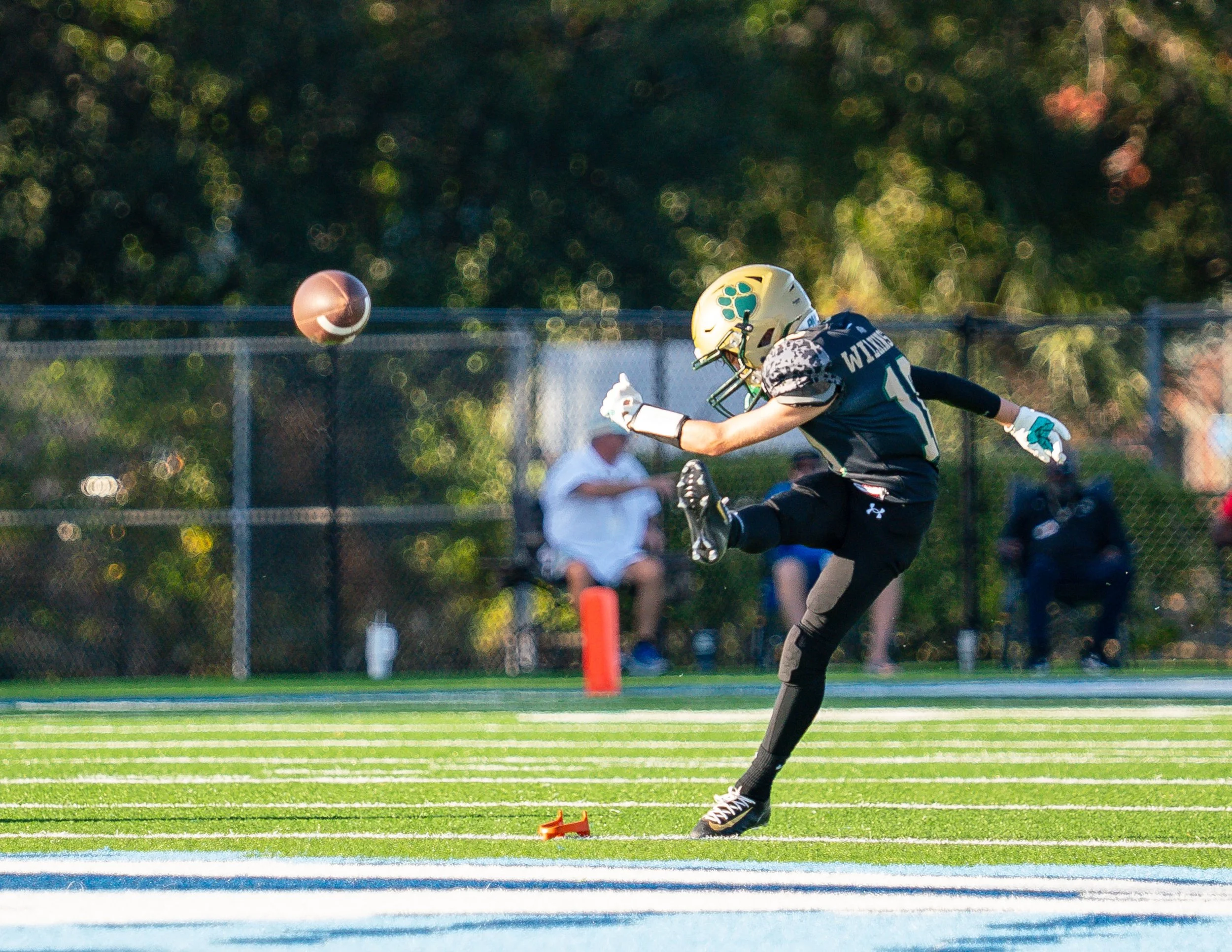A football player in a black uniform and yellow helmet making a kick on a football field during a game.