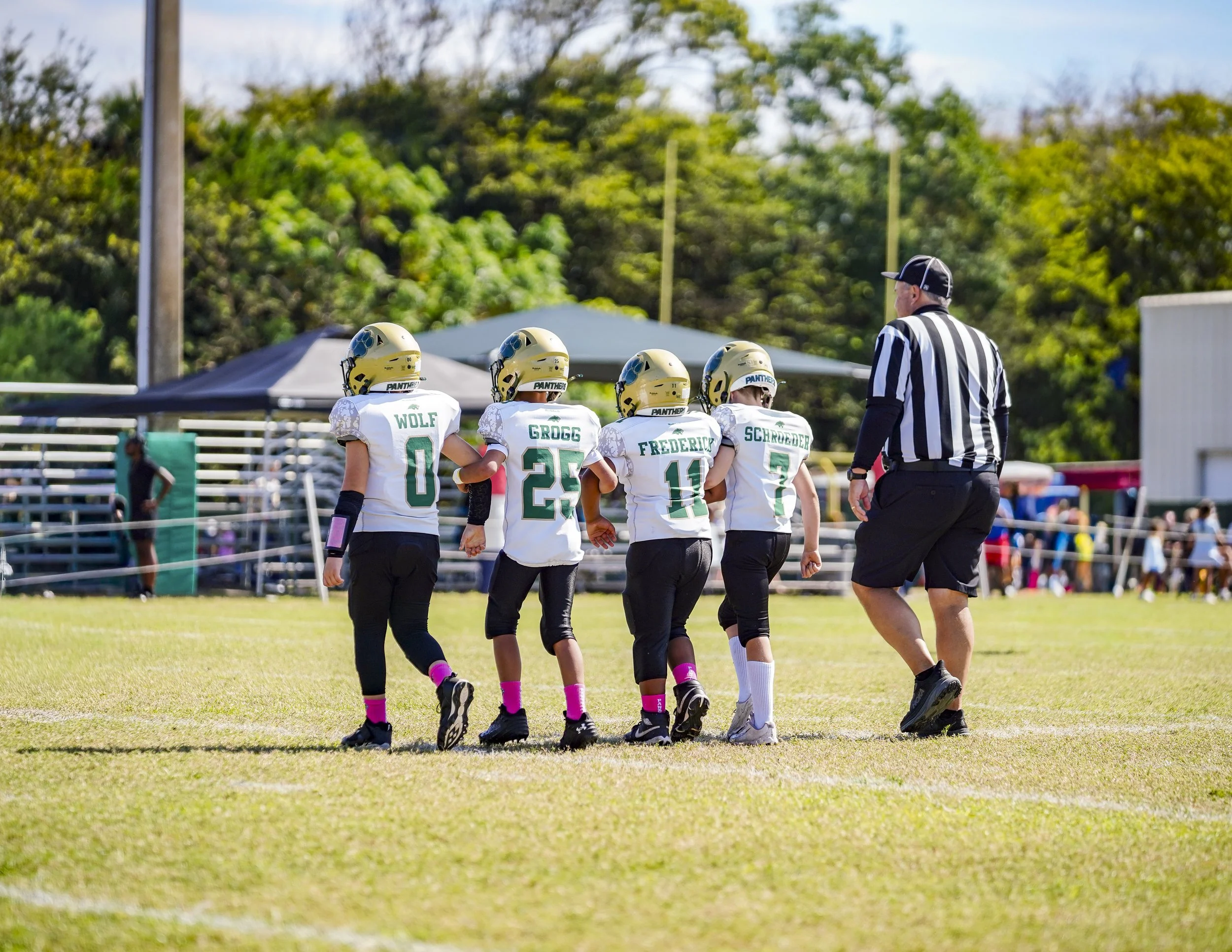 Four young football players wearing white jerseys, black pants, and gold helmets walking on a football field with a coach or referee in black shorts and a striped shirt nearby. Spectators are visible in the background.