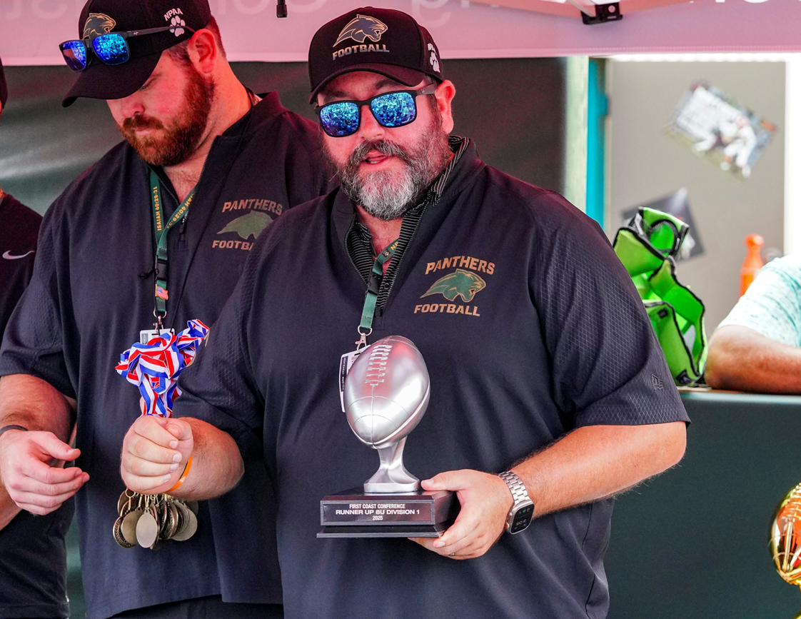 A man wearing sunglasses, a cap, and a black shirt with 'Panthers Football' logo is holding a trophy shaped like a football. He is also holding medals in his left hand. Another person next to him is partially visible, also wearing a 
