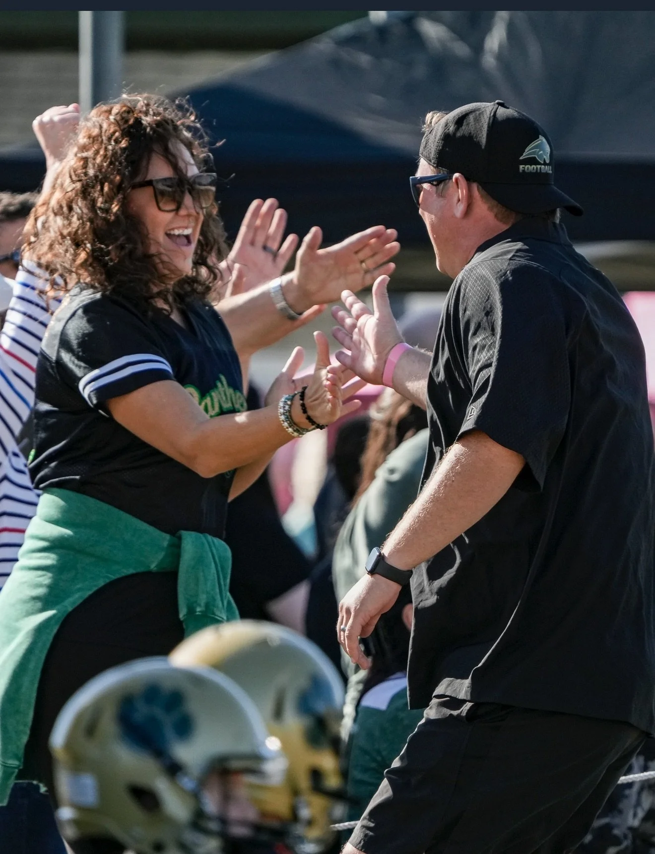 People celebrating at a football game, smiling and high-fiving, with football helmets on the ground in the foreground.
