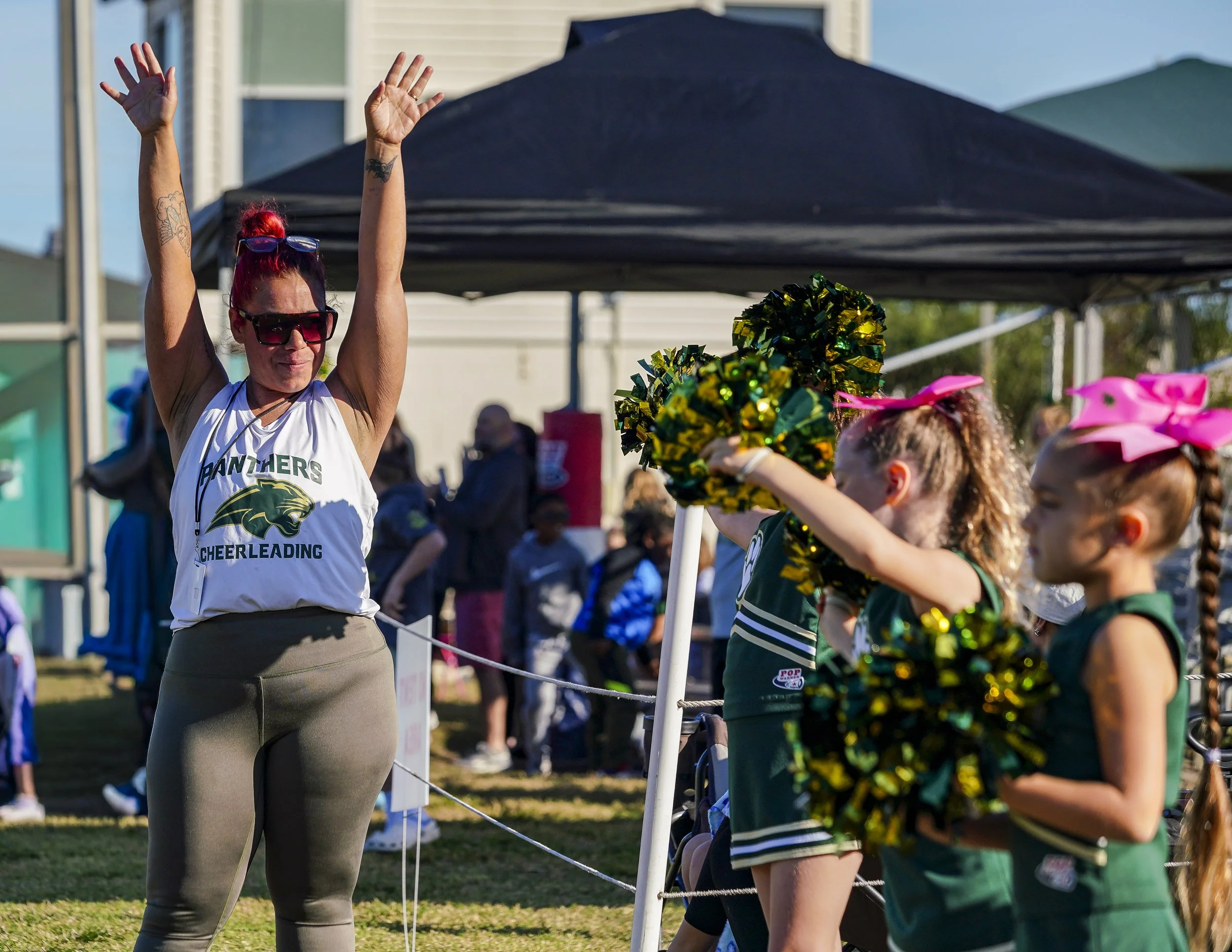 A cheerleader stands with arms raised, wearing sunglasses and a white cheerleading shirt with a panther logo, while young cheerleaders in green uniforms with pink bows cheer with pom-poms during an outdoor event.