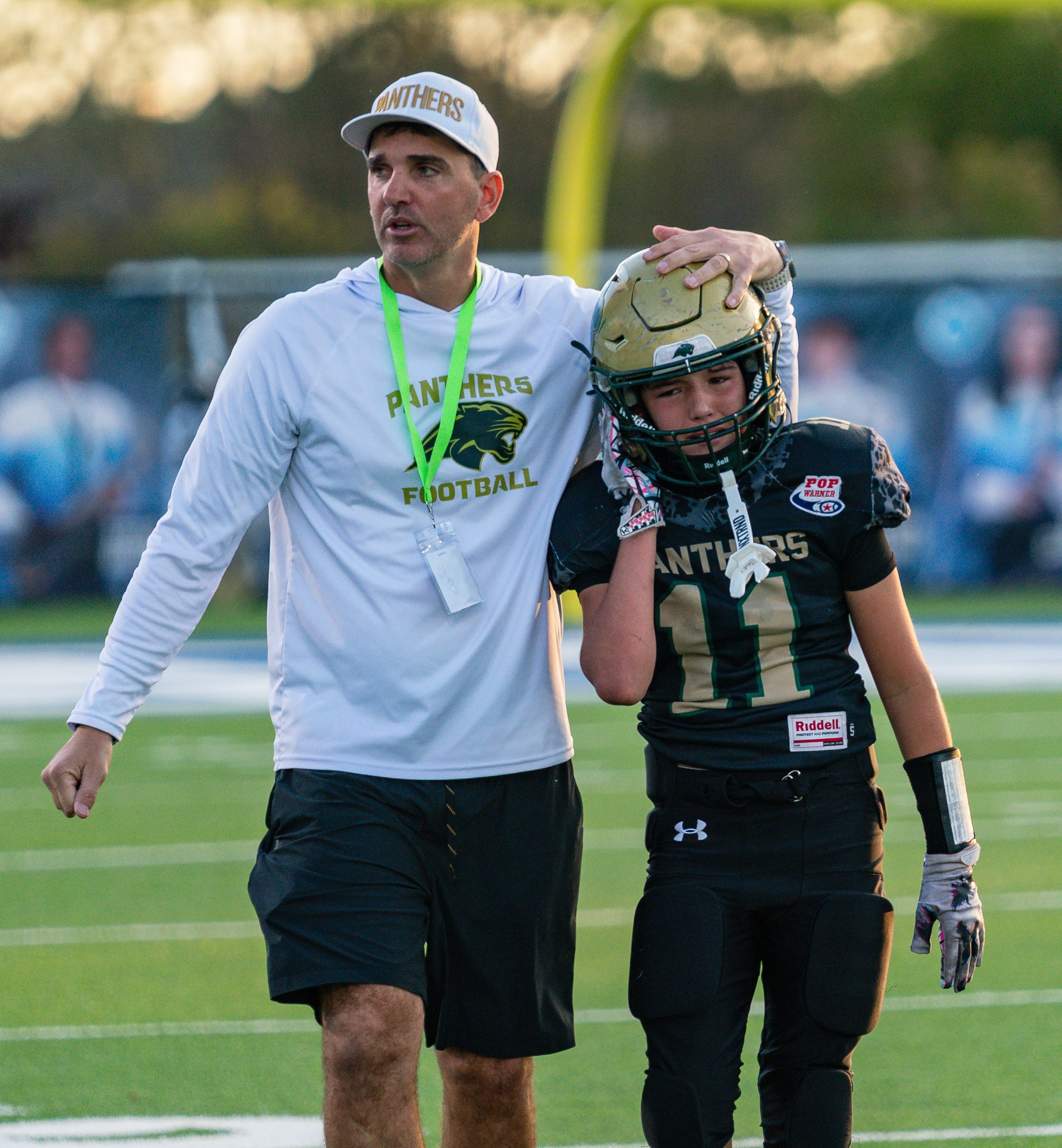 A football coach with a panther logo on his shirt walking on the field alongside a female football player holding her helmet, both dressed in black and gold, during a game.