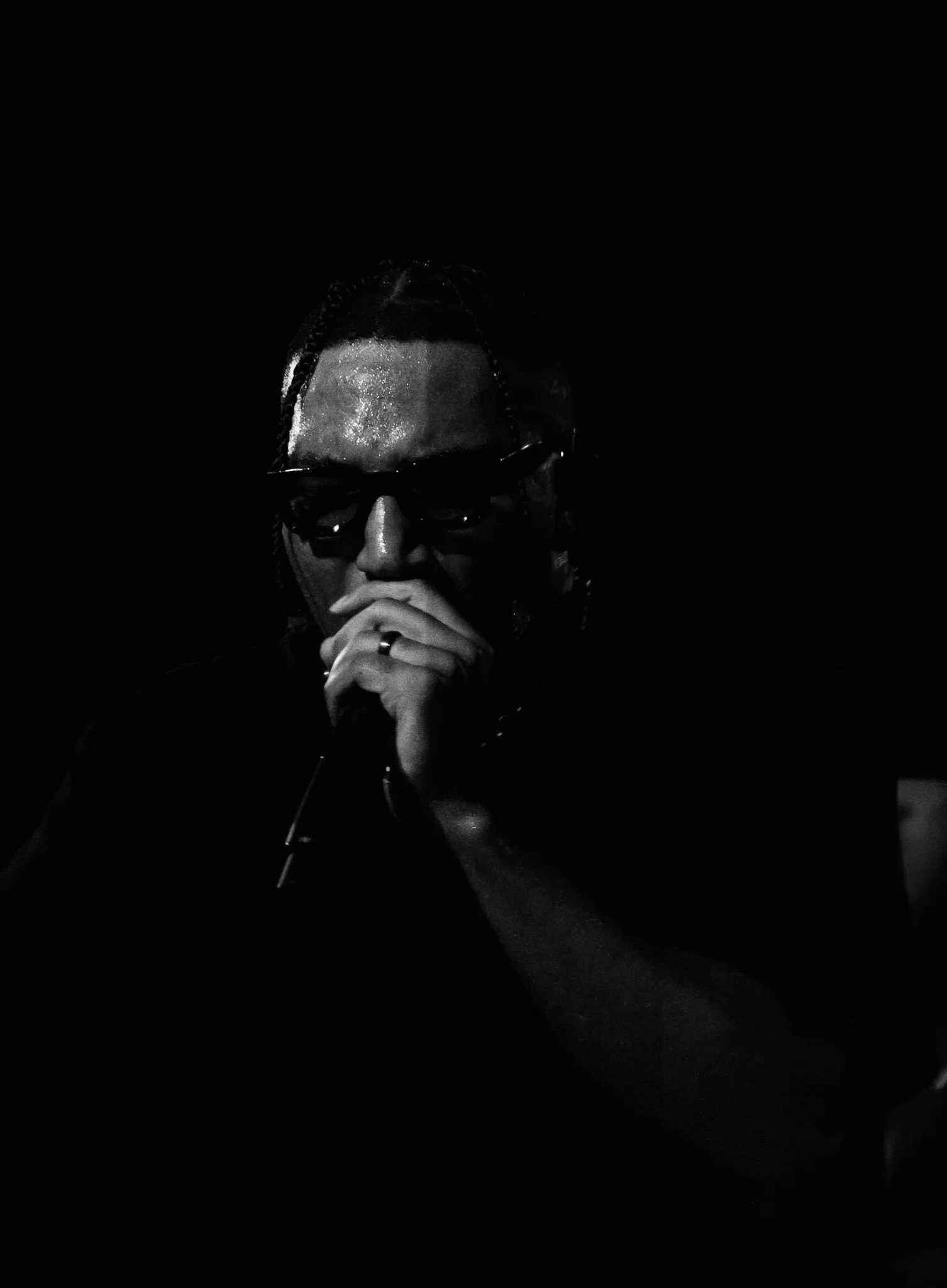 A person with braided hair and glasses holds a microphone close to their mouth in a dark setting, with beads of sweat visible on their forehead.