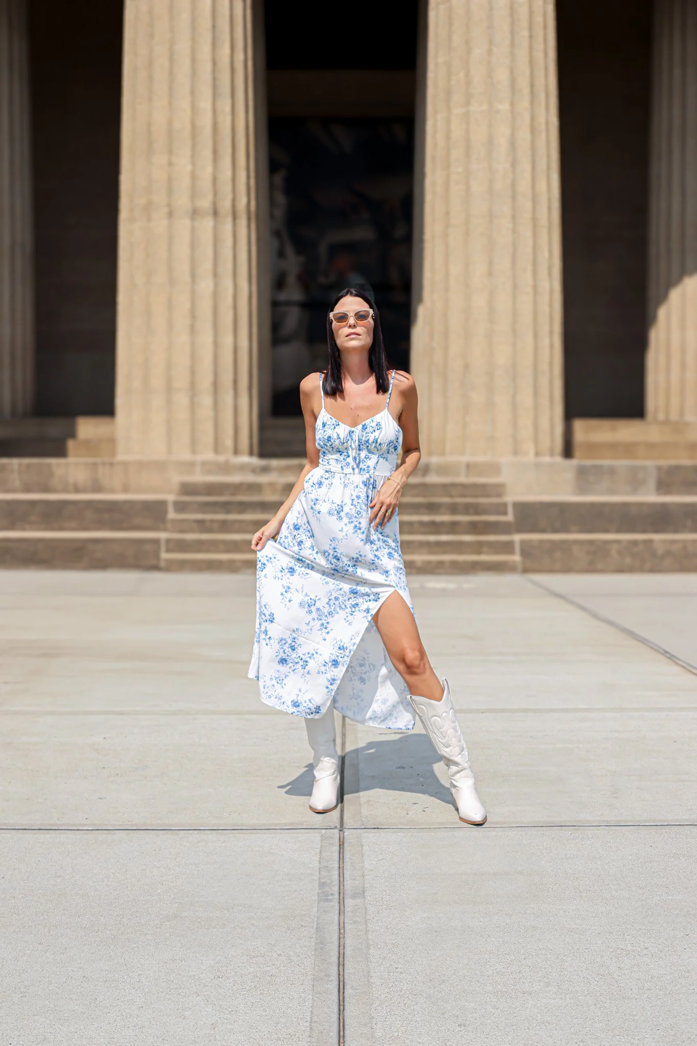 A woman in a white and blue floral dress with white cowboy boots standing in front of large ancient Greek-style columns.