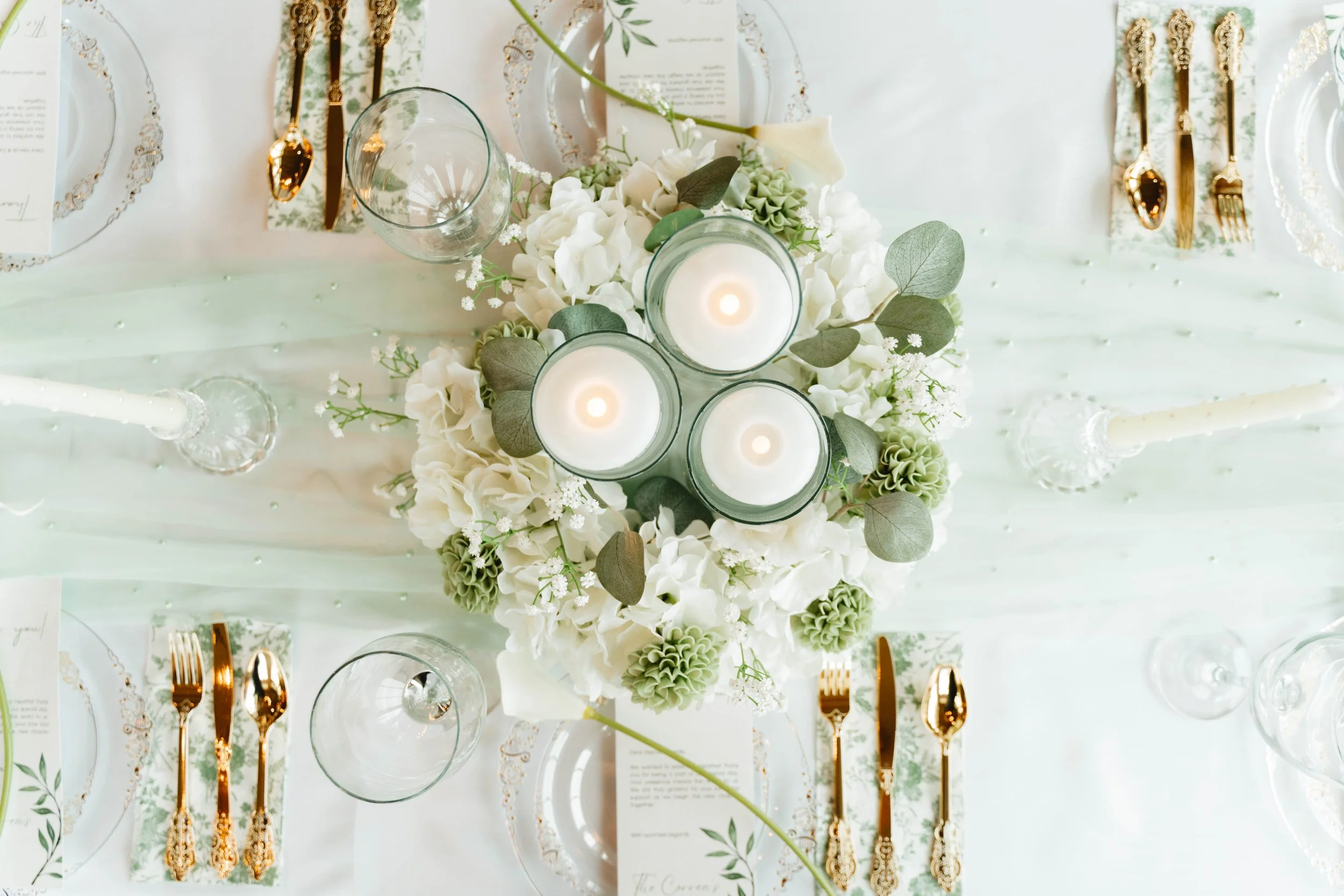 Elegant table setting with a floral centerpiece featuring white and green flowers, three lit candles in glass holders, gold cutlery, and clear glassware on a white tablecloth.