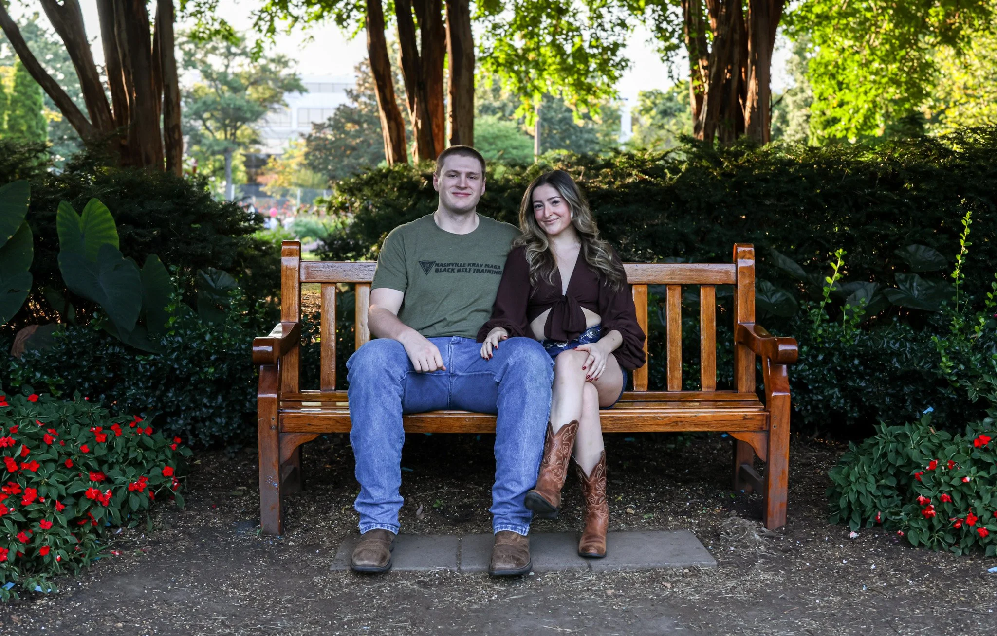 A young man and woman sitting on a wooden park bench surrounded by greenery and red flowers, with trees and a park in the background.