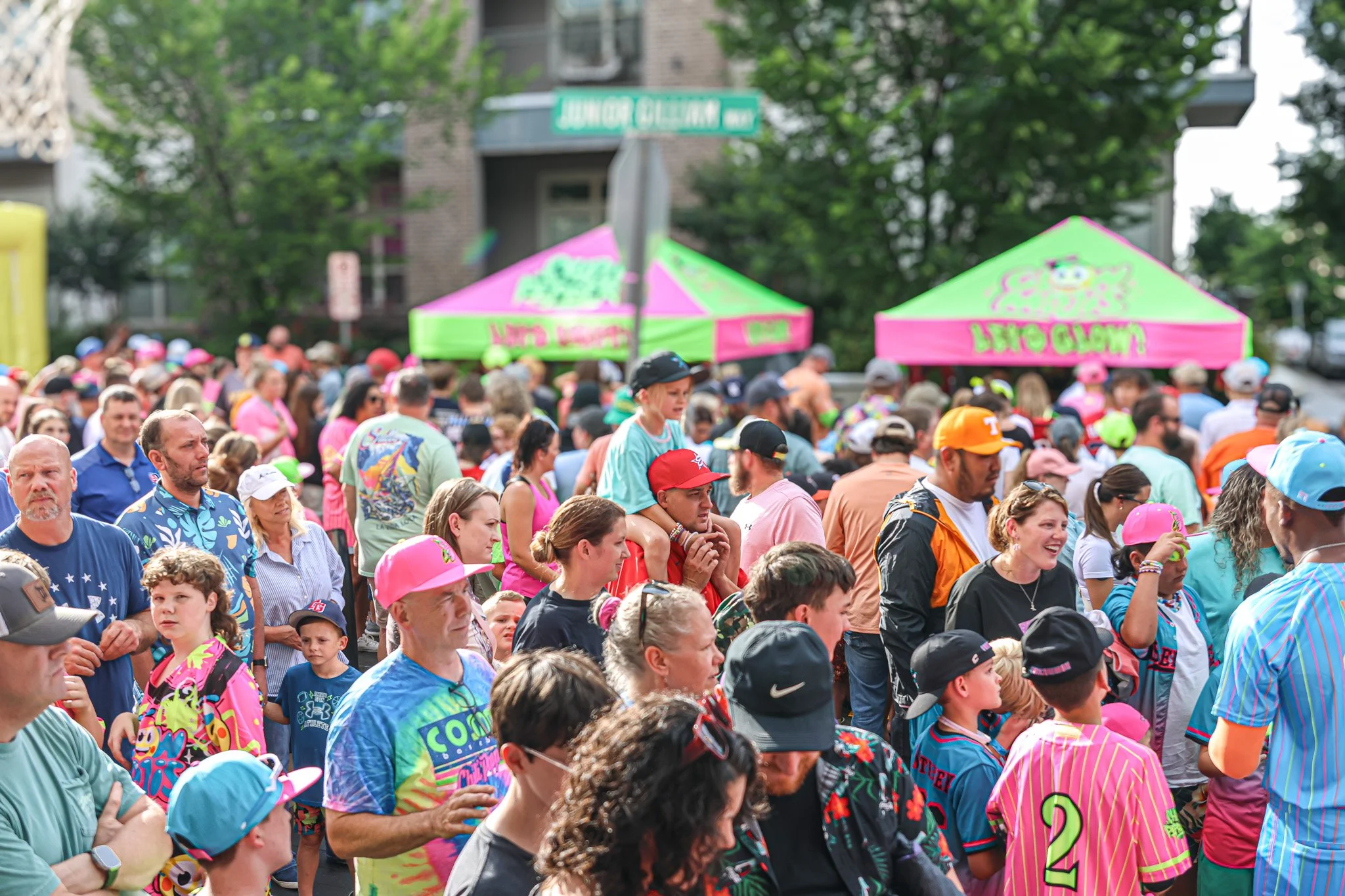 A crowd of people gathered outdoors for a community event with pink and green tents in the background, trees, and a street sign that reads 'Junior Gillum Way'.