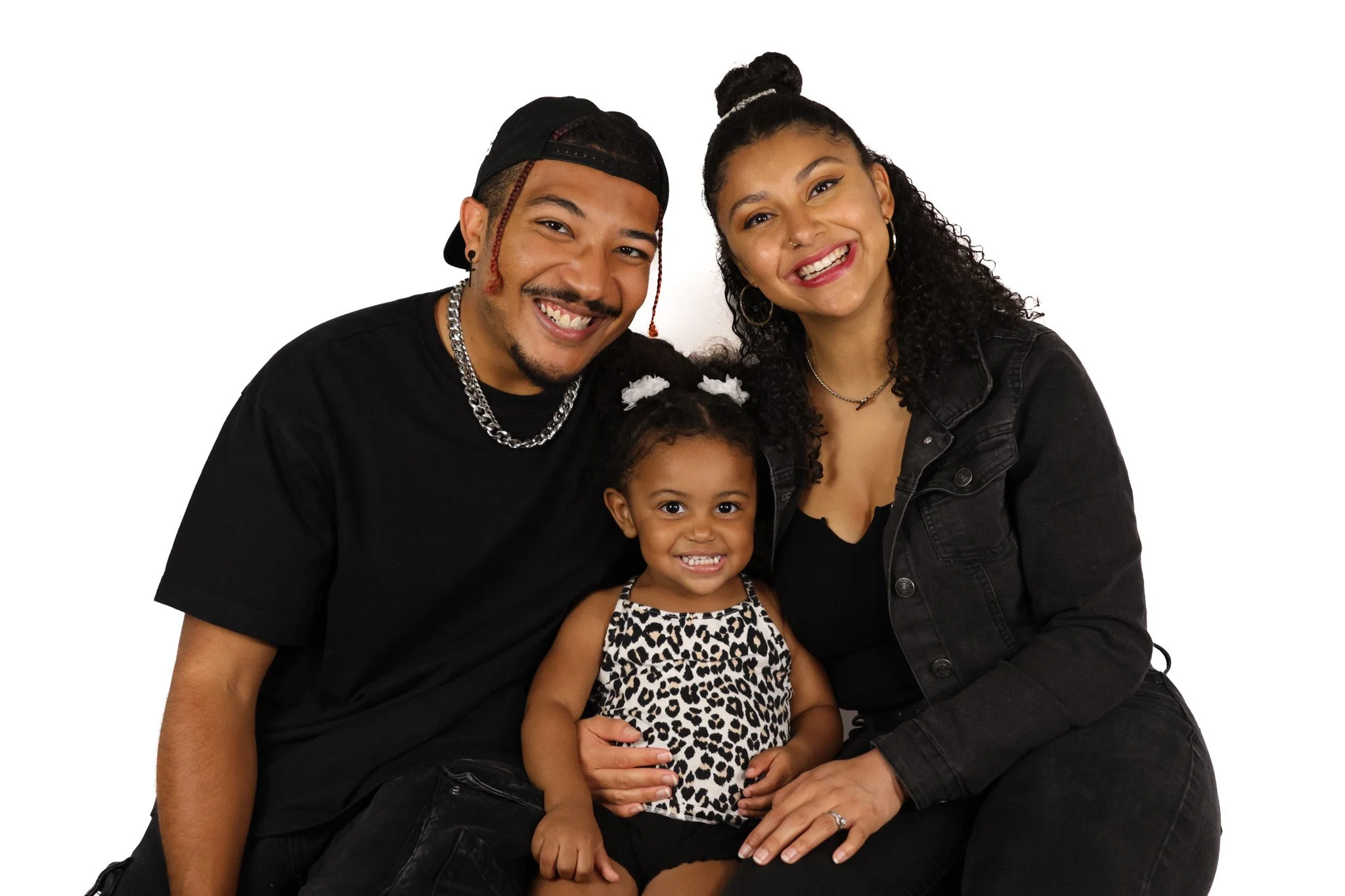 A happy family of three smiling and sitting together against a white background. The father has a beard, mustache, and is wearing a black cap, black shirt, and silver jewelry. The mother has curly hair, earrings, and is dressed in a black top and bla