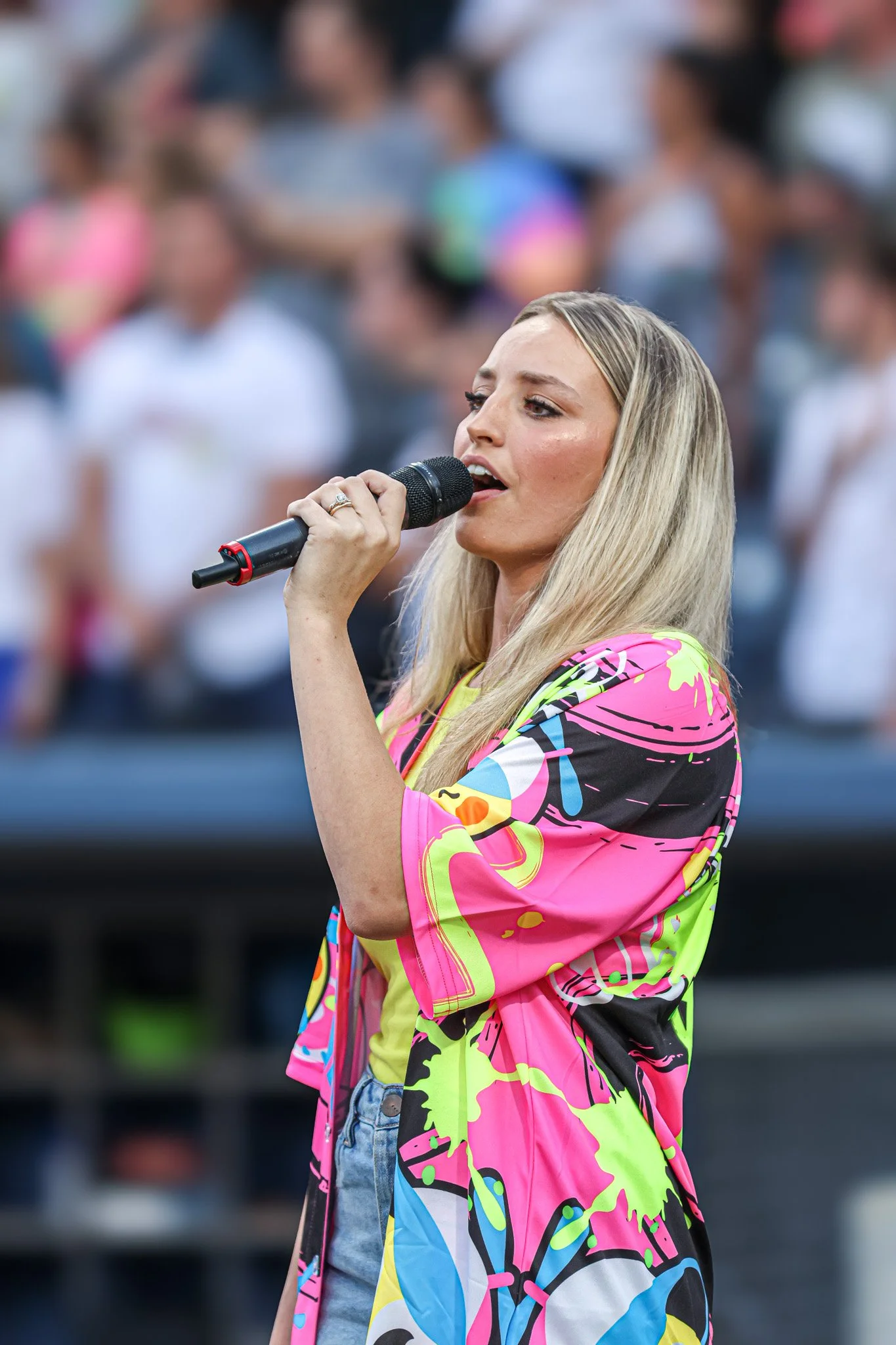 A young woman with long blonde hair singing into a microphone at an outdoor event. She is wearing a colorful, graffiti-style jacket and a yellow shirt, with a serious expression on her face. Blurred crowd in the background.