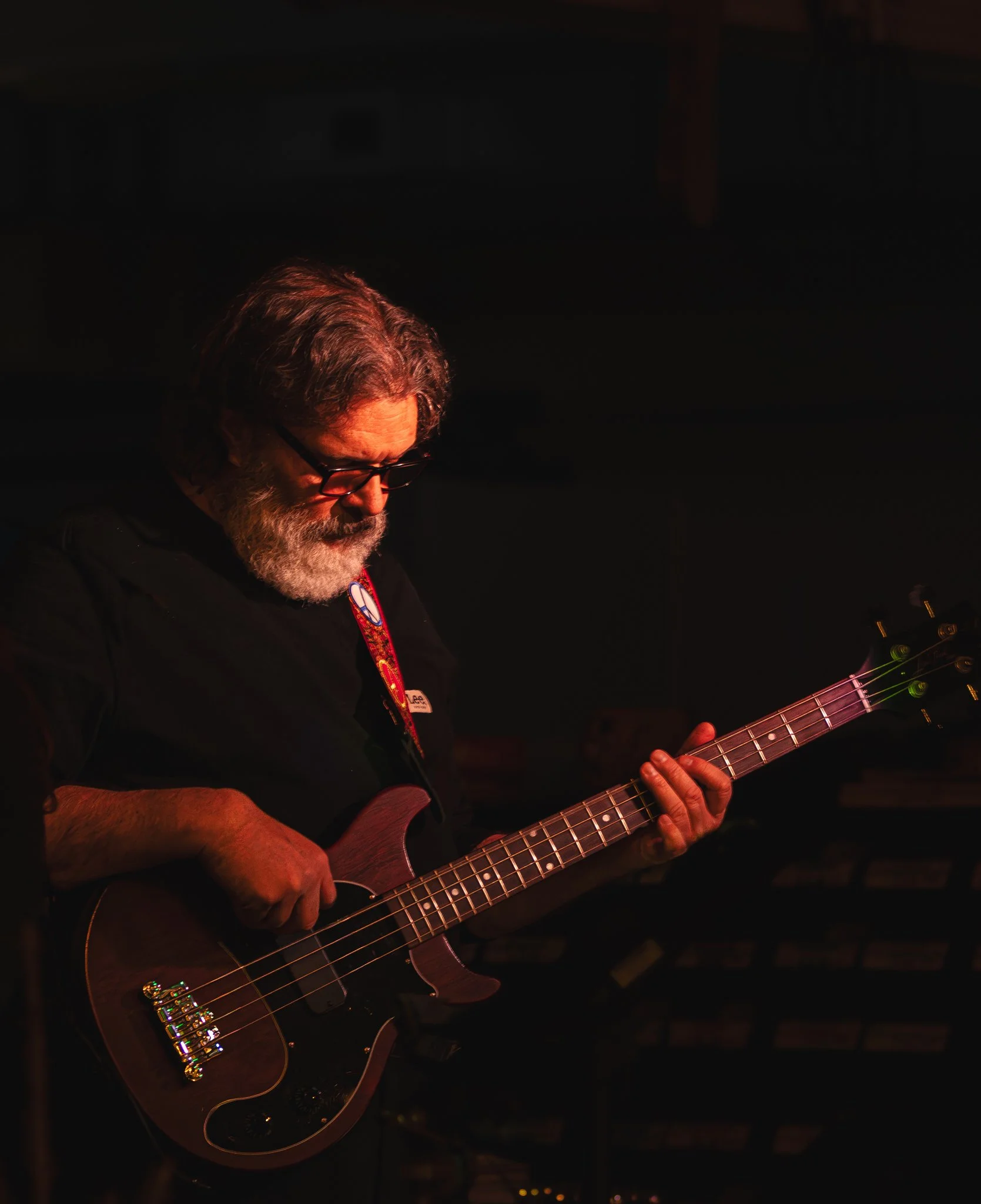 A bearded man with glasses playing an electric bass guitar on stage in low light.