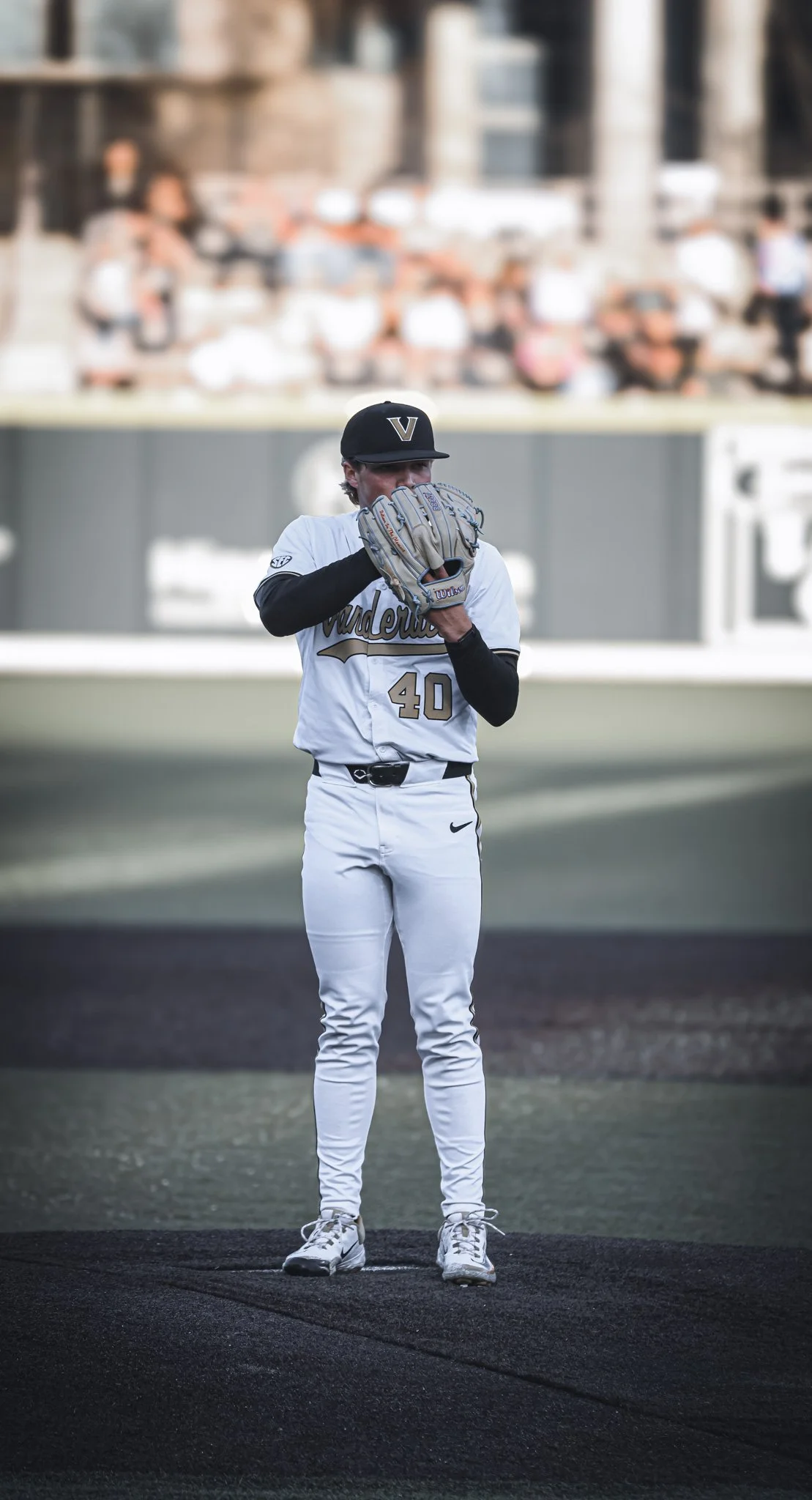 A baseball player standing on the mound in uniform, wearing a black cap with a 'V', holding a baseball glove over his face, in front of an outfield with spectators.