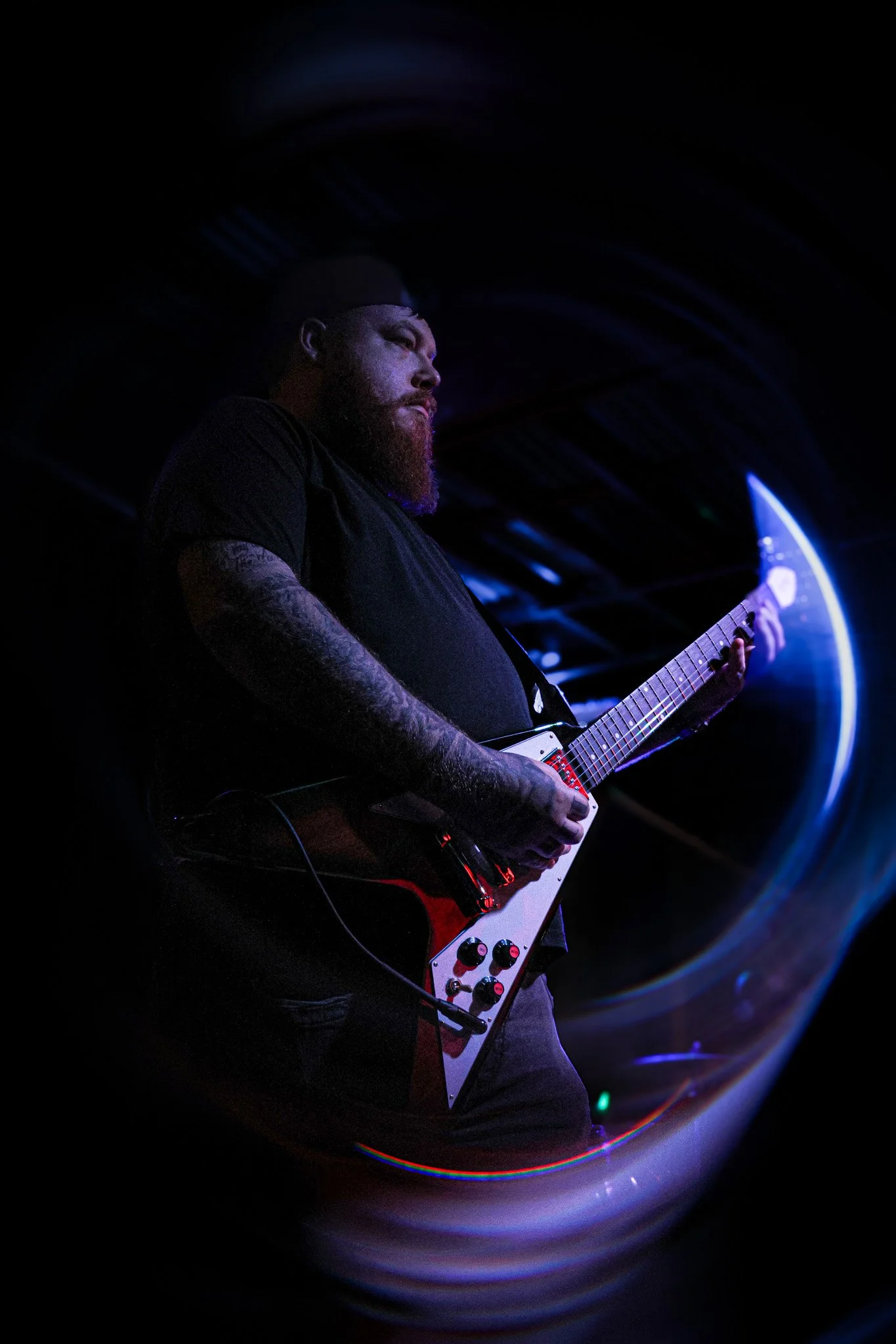 A man with a beard and tattooed arms playing an electric guitar on stage with blue and purple lighting.